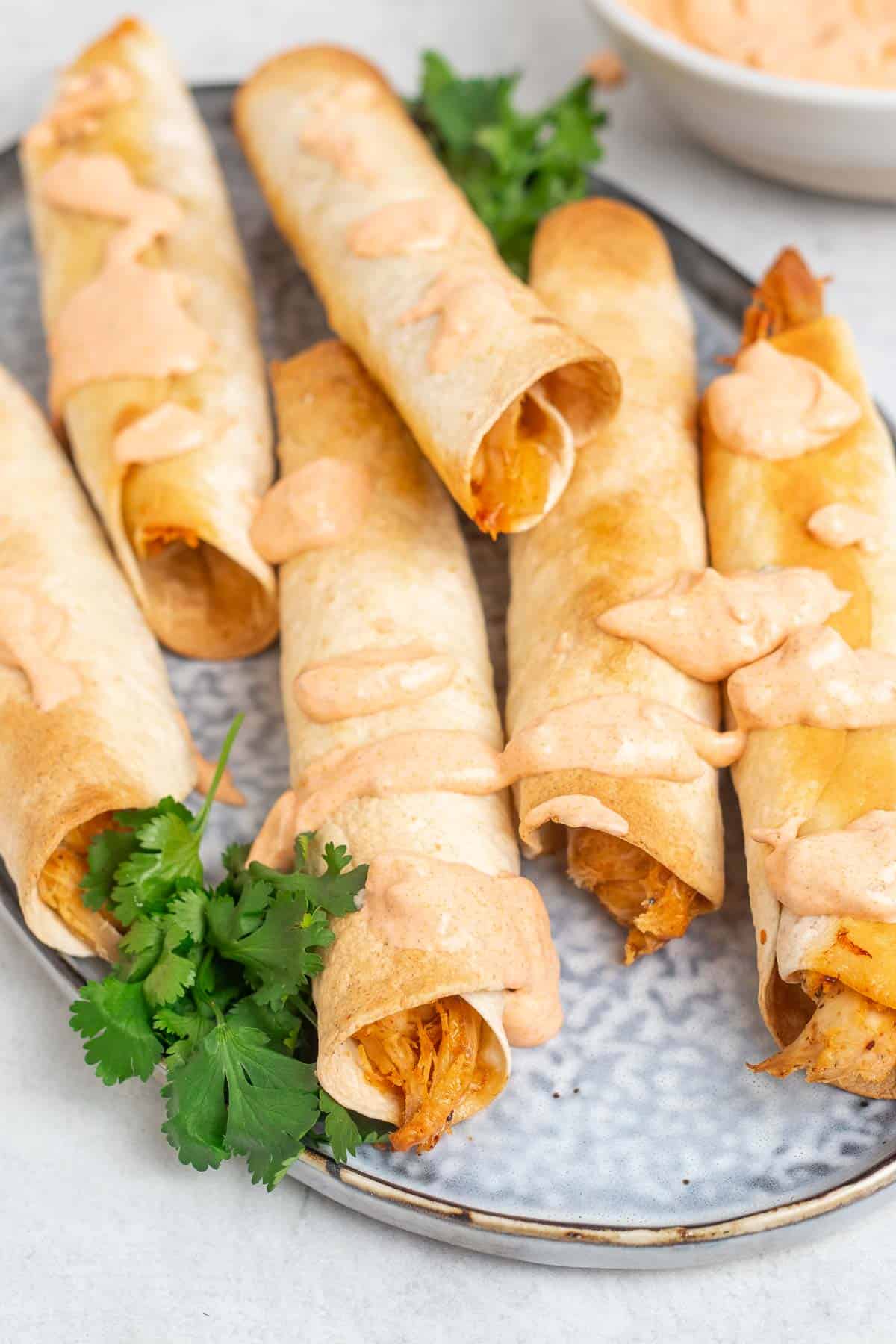 Close up of six taquitos on a plate with fresh cilantro and drizzled with sauce