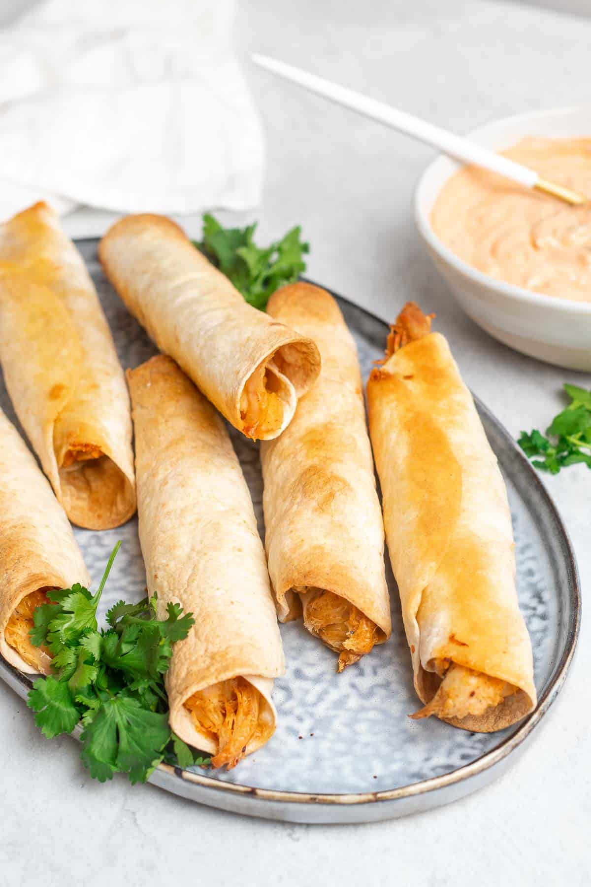 Six taquitos on a plate with fresh cilantro next to a white ramekin of sauce