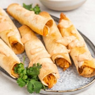 Six baked chicken taquitos on a plate, drizzled with sauce and served with fresh cilantro