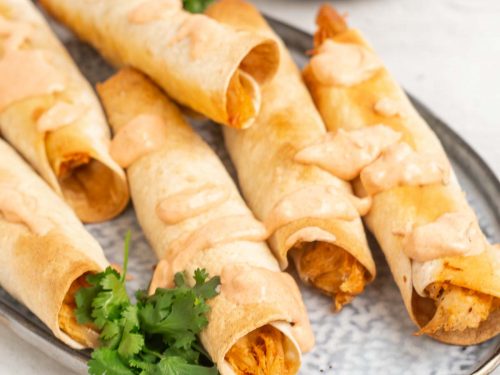 Six baked chicken taquitos on a plate, drizzled with sauce and served with fresh cilantro