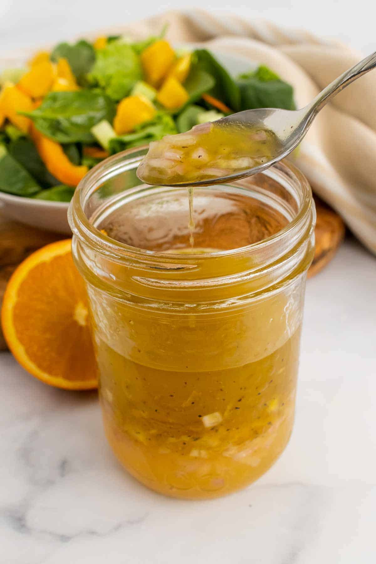 Spoon with homemade citrus vinaigrette above glass jar in front of salad