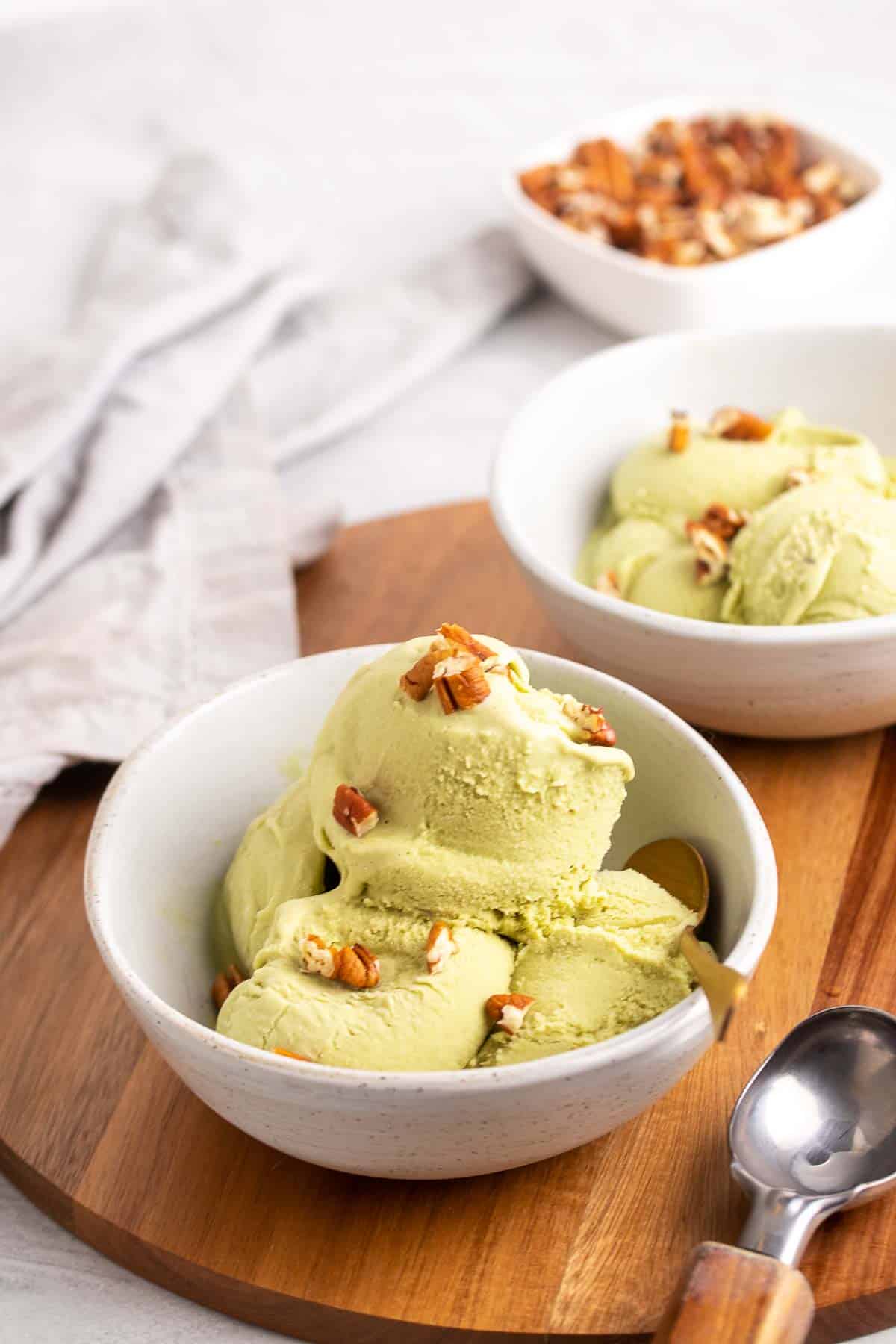 Two white bowls , each with several scoops of green ice cream inside, topped with chopped pecans, on top of a wooden serving tray