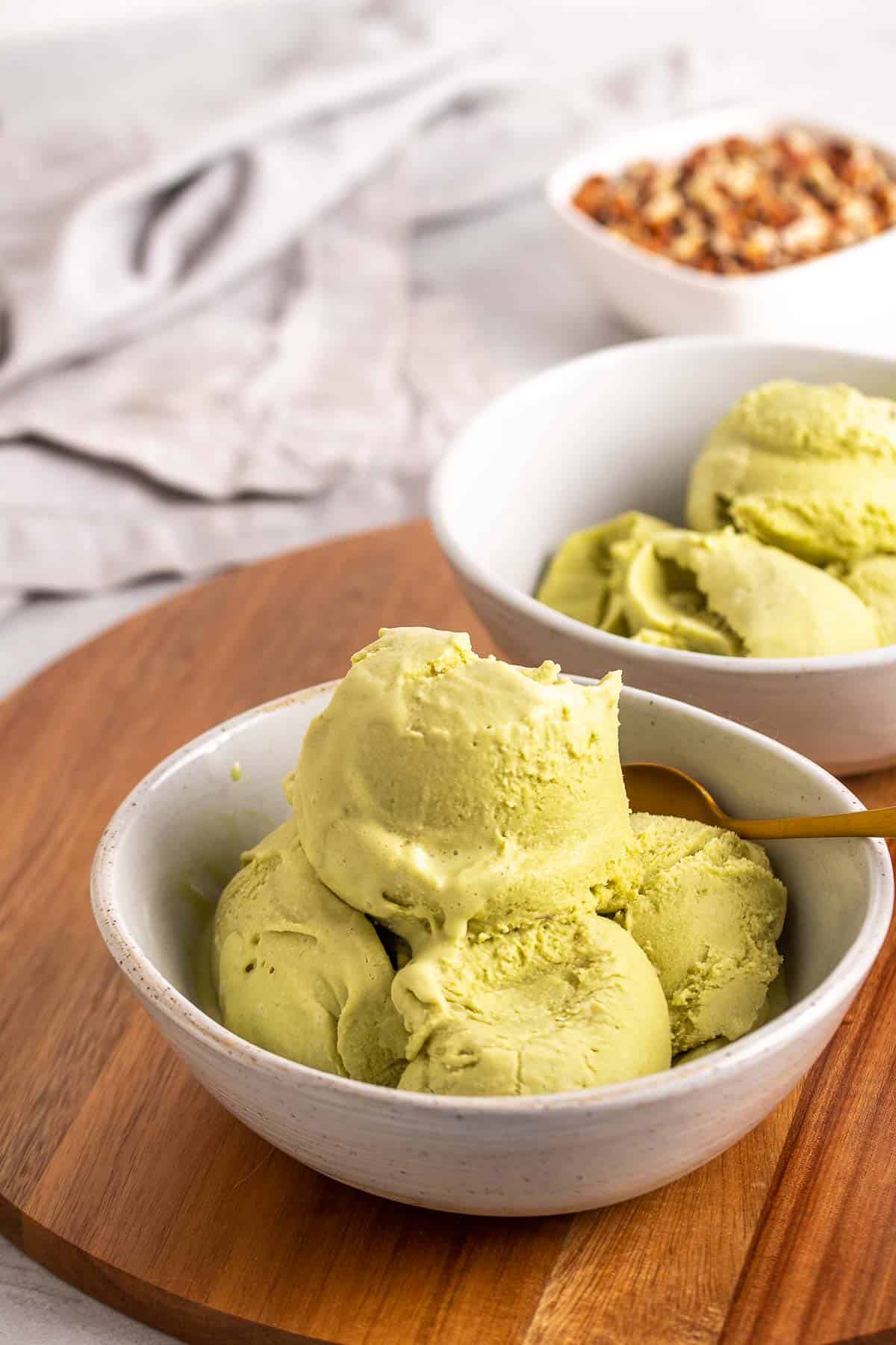 Four scoops of matcha green tea ice cream in a white bowl with a second bowl in the background, both on a wooden serving board