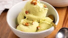 Four scoops of matcha green tea ice cream in a white bowl with a gold spoon, topped with chopped pecans, on a wooden serving tray
