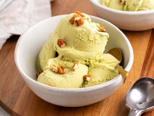 Four scoops of matcha green tea ice cream in a white bowl with a gold spoon, topped with chopped pecans, on a wooden serving tray