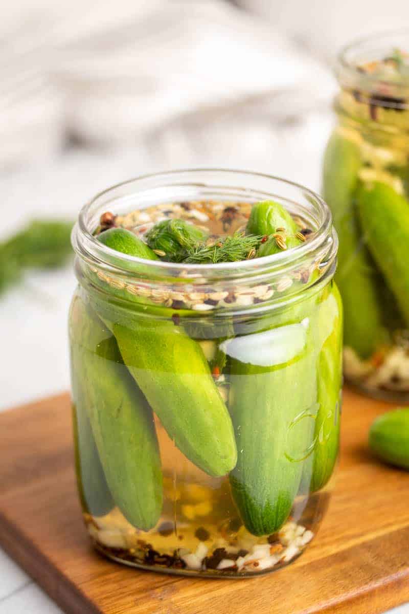 A glass jar of cucumbers in the pickling mixture on a wooden serving tray