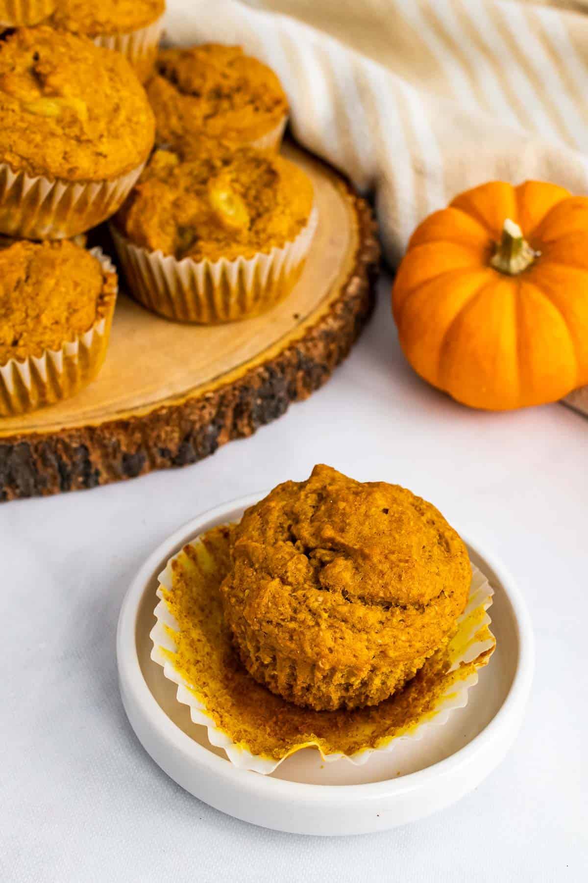 One muffin on a small white plate in front of a serving tray with more muffins; pumpkin in the background