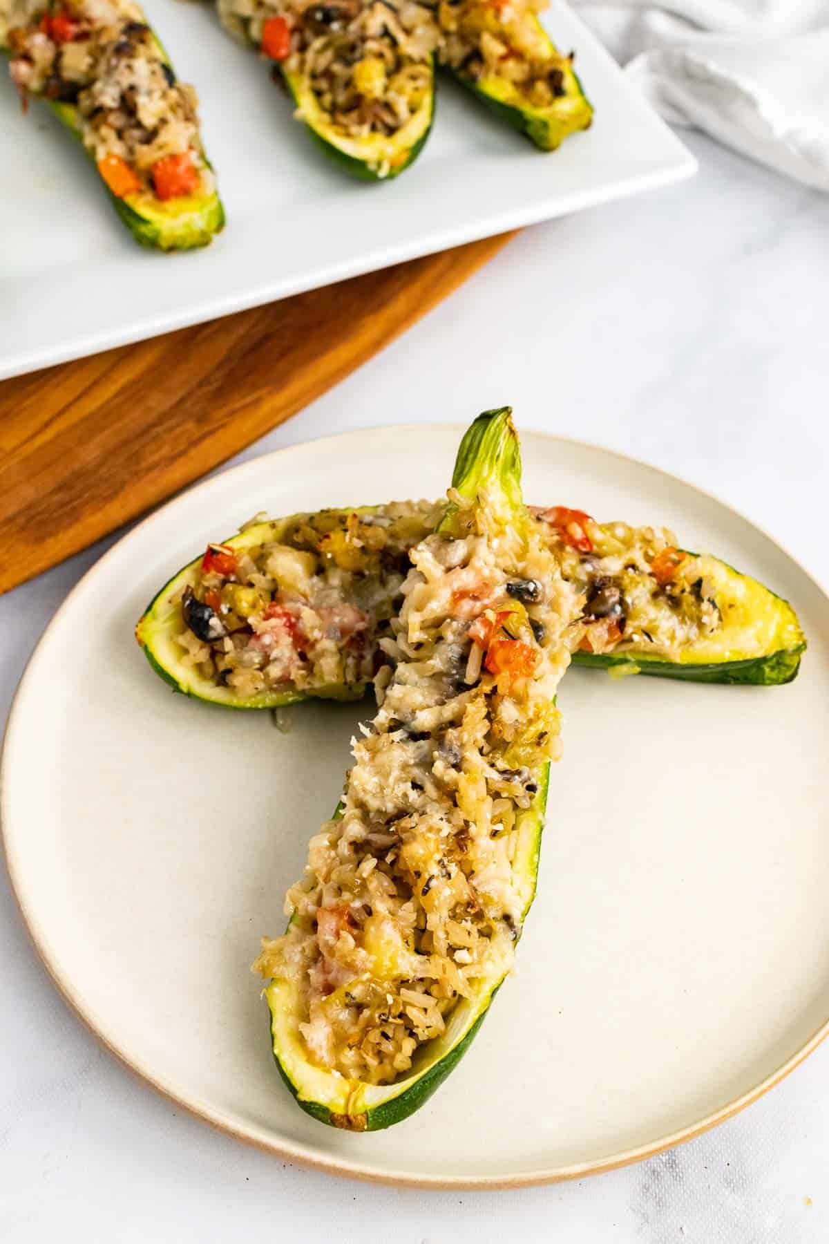 Two vegetarian stuffed zucchini halves on a white plate with a platter in the background