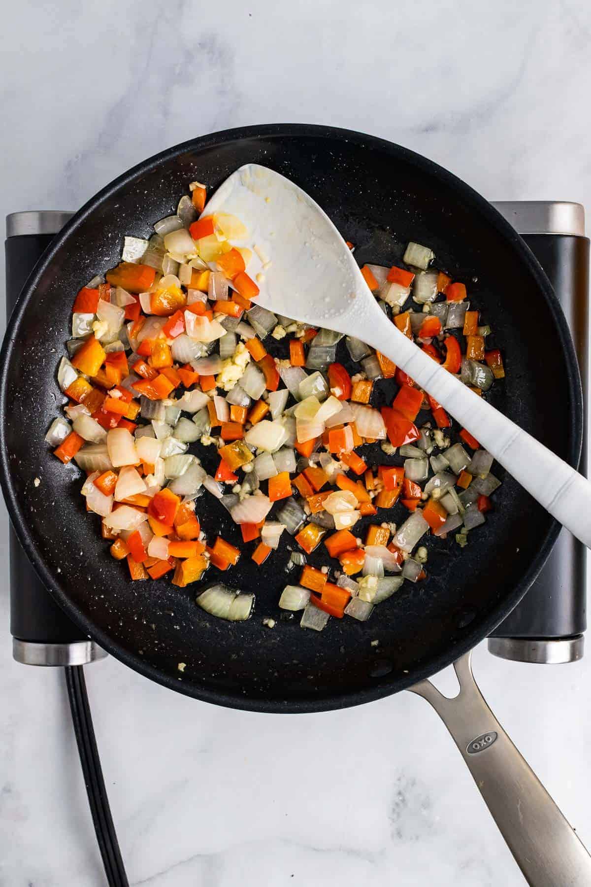 Diced onions and peppers cooking in a pan in oil with a spatula