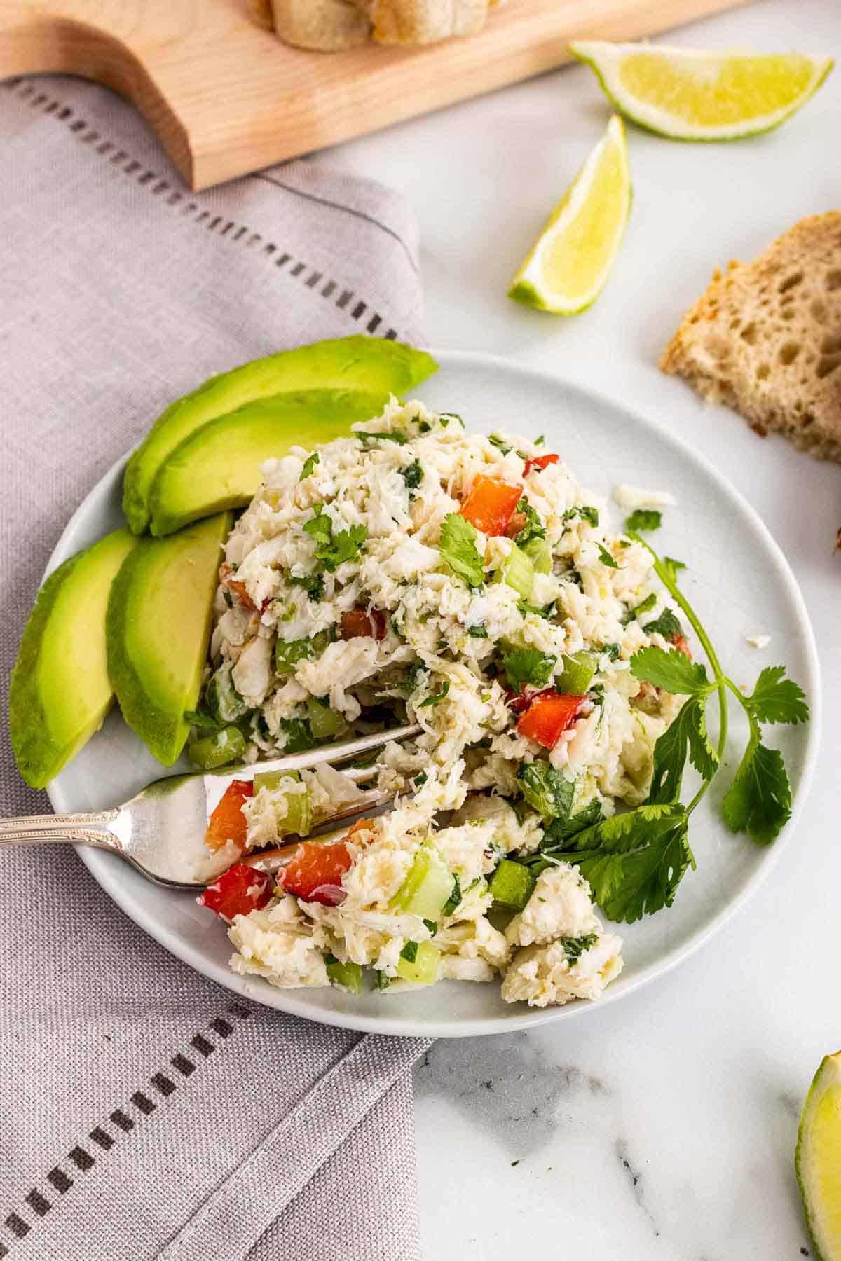Crab mix on a white plate with avocado slices and garnished with a parsley sprig next to slices of toast and lime wedges