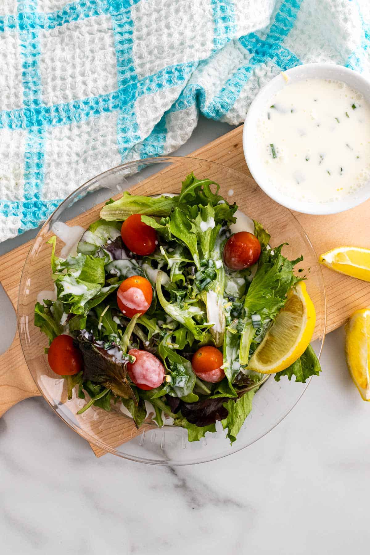 Dressing in a white ramekin on a wooden serving tray next to a wedge of lemon and a fresh salad with dressing on top, as seen from above