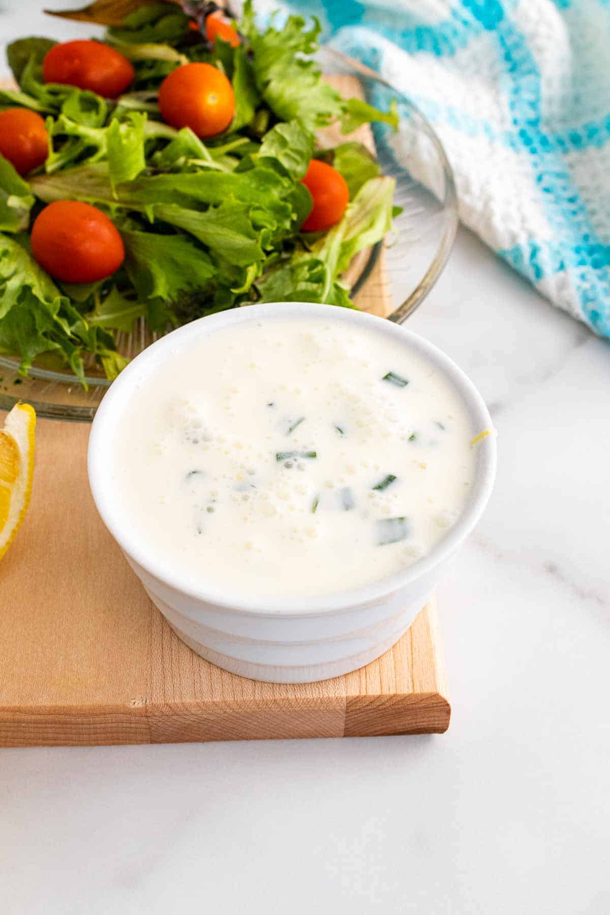 Creamy Lemon-Chive Dressing in a white ramekin on a wooden serving tray next to a wedge of lemon and a fresh salad with dressing on top