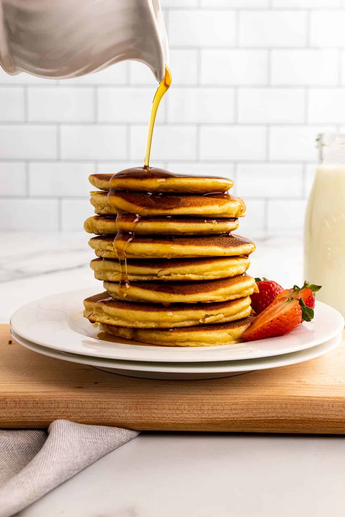Stack of pancakes on a plate with strawberries, syrup being poured on top from a white ramekin