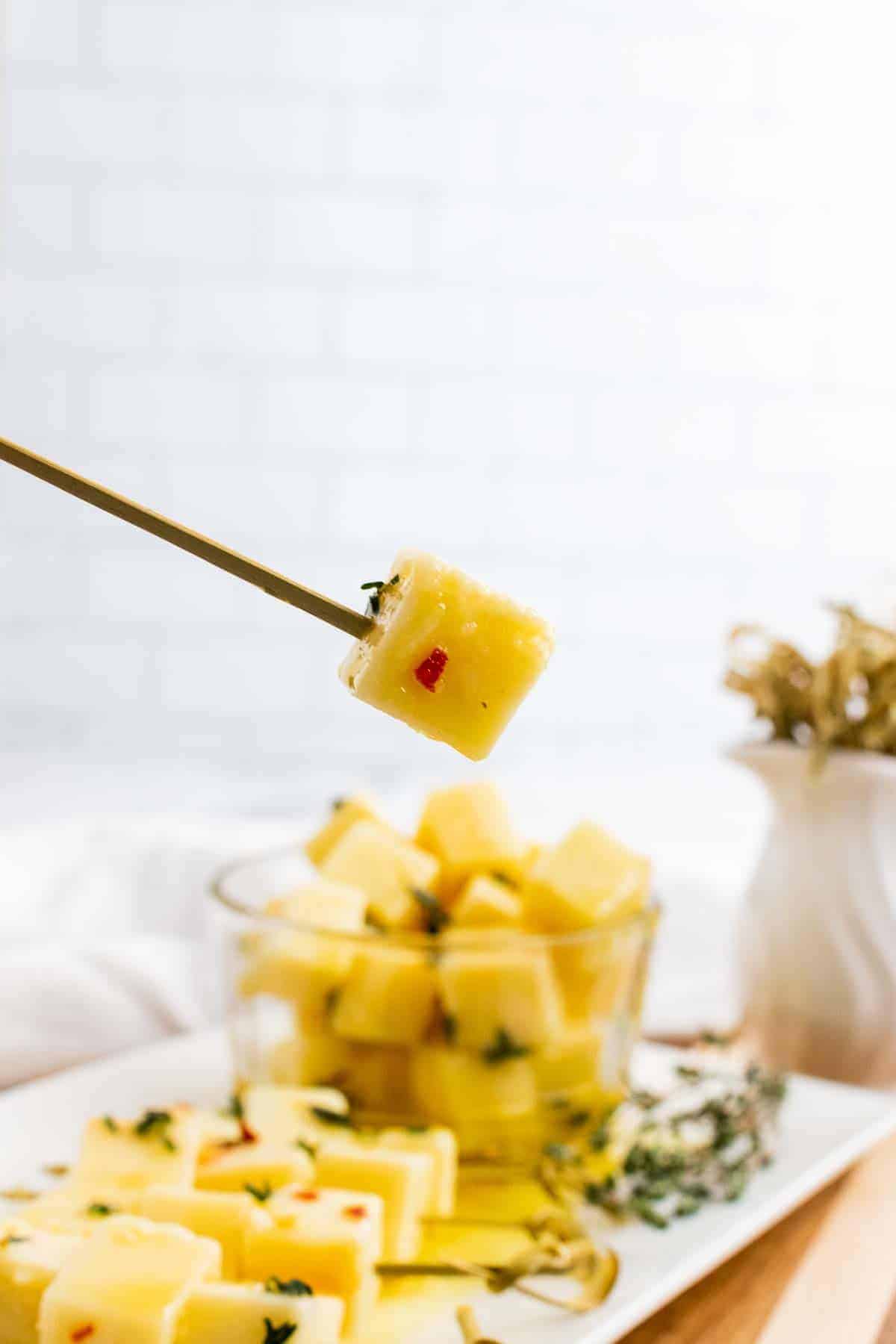 A cube of honey-thyme marinated cheddar cheese on a skewer over a serving tray with more cheese skewers and a bowl of cheese in the background