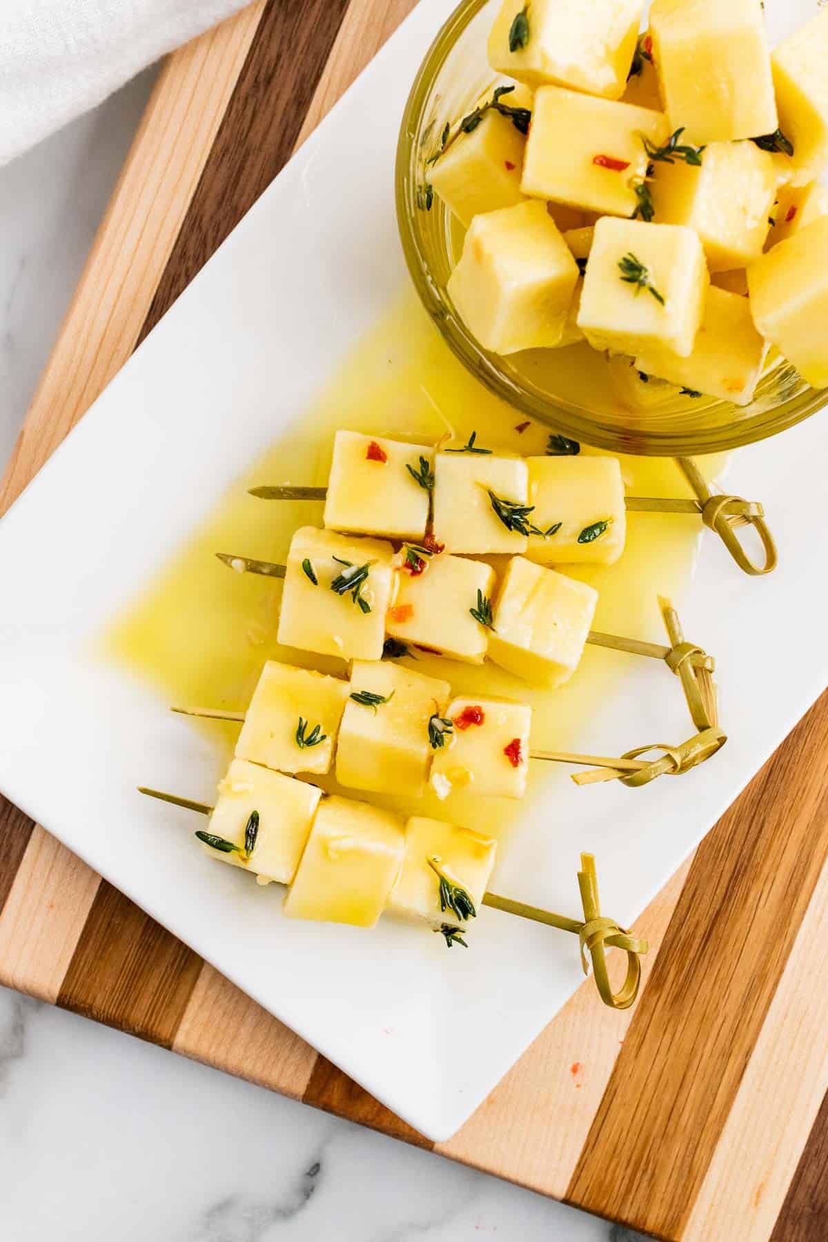 Skewers with three cheese cubes each next to a ramekin of more cheese cubes on a wooden serving tray