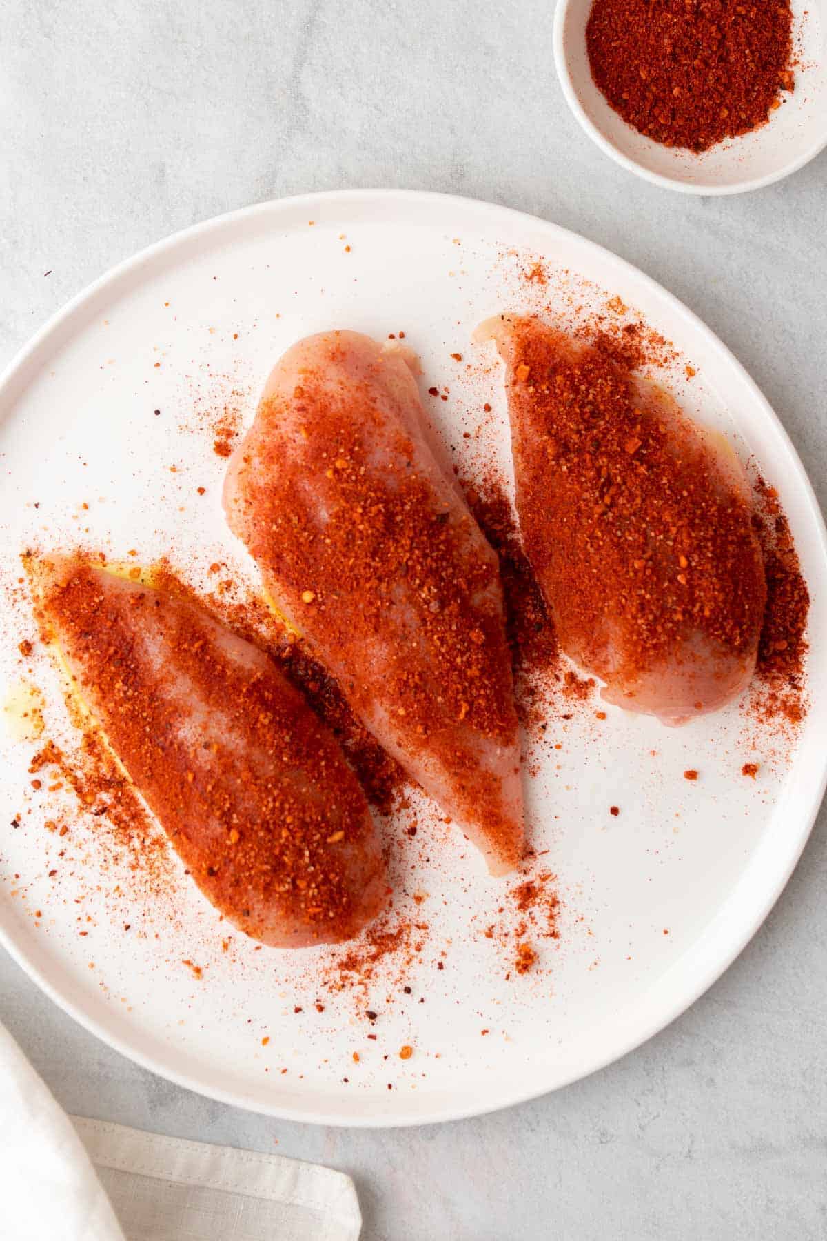 Three raw chicken breasts on a white plate seasoned with taco seasoning, as seen from above