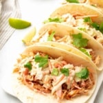 Close-up of low sodium chicken tacos in a row of three topped with cilantro on a white plate next to lime wedges
