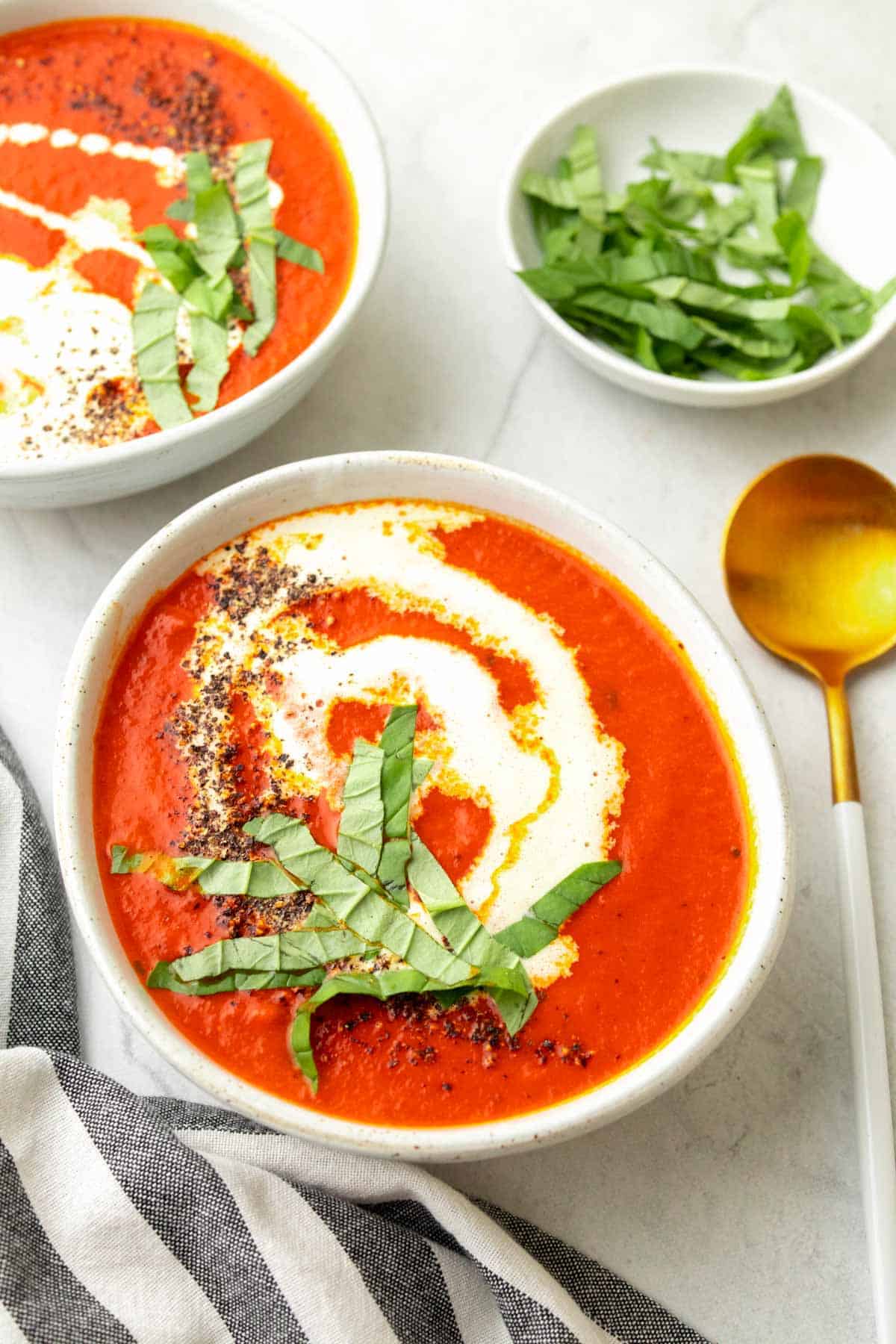 Low sodium tomato soup in a white bowl with cream, fresh basil leaves, and black pepper on top next to a gold spoon and cloth napkin