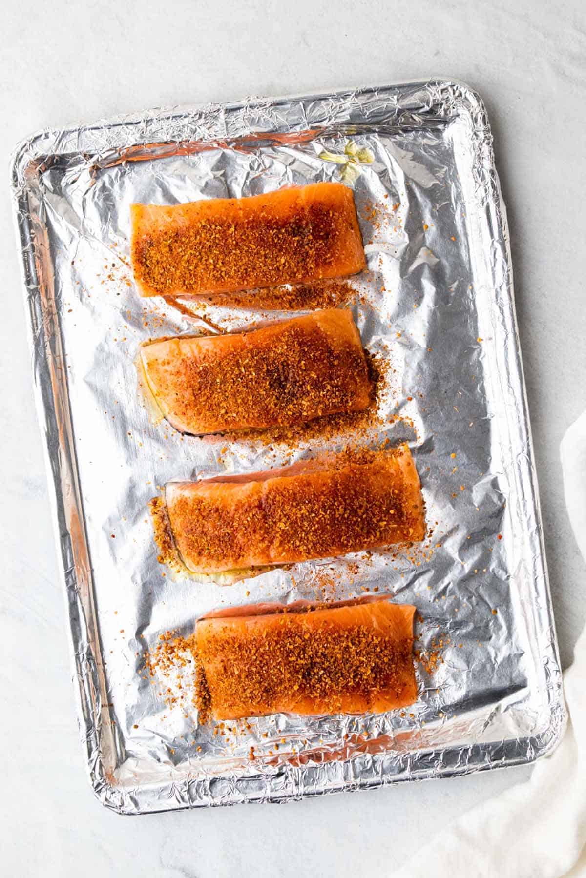 Four salmon fillets topped with olive oil and taco seasoning on a baking sheet lined with aluminum foil, as seen from above