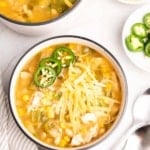 Two bowls of low sodium white chicken chili topped with jalapeno slices and cheese next to ramekins with more toppings and two silver spoons, as seen from above
