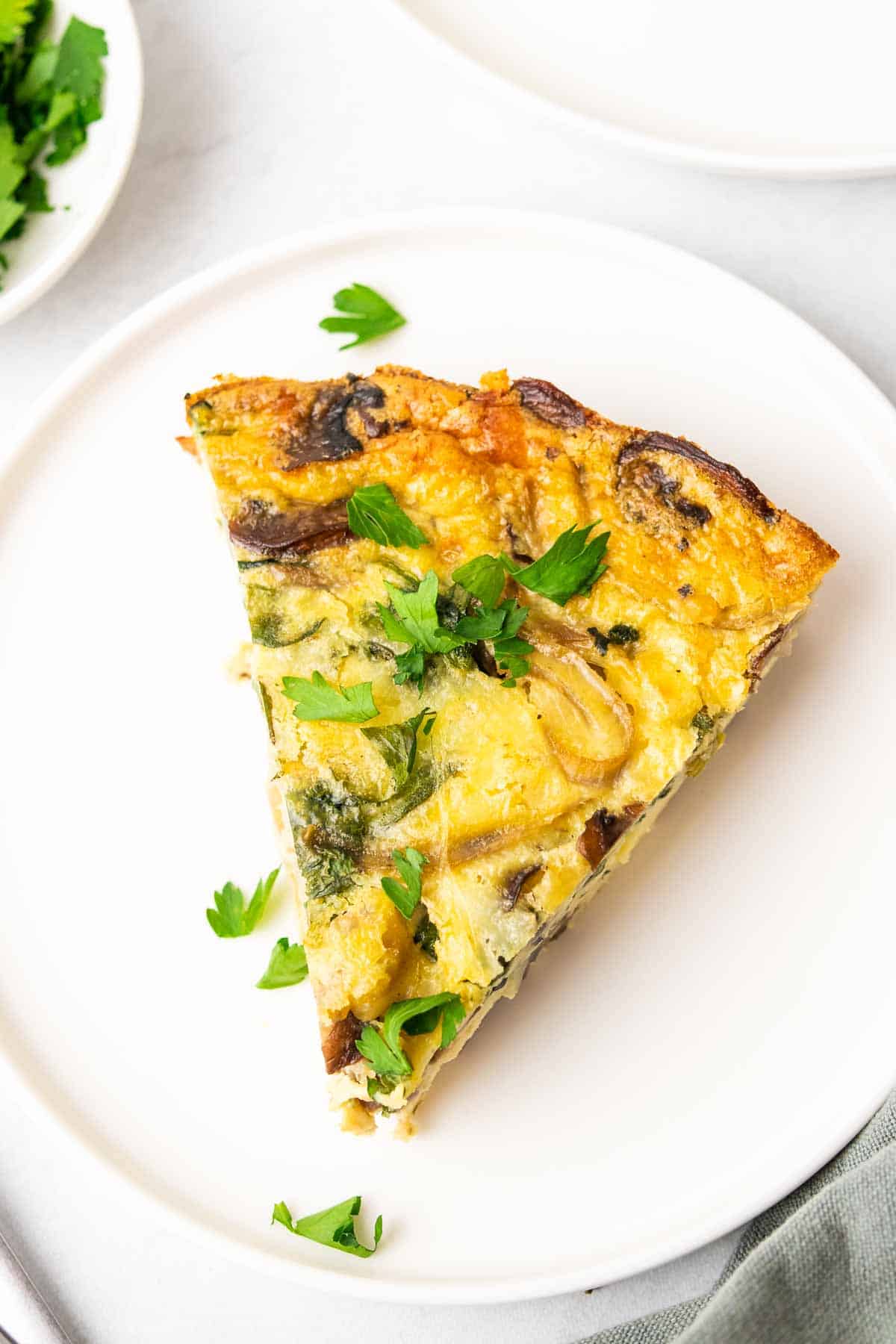Closeup of a slice of low sodium spinach quiche on a white plate with fresh herbs on top