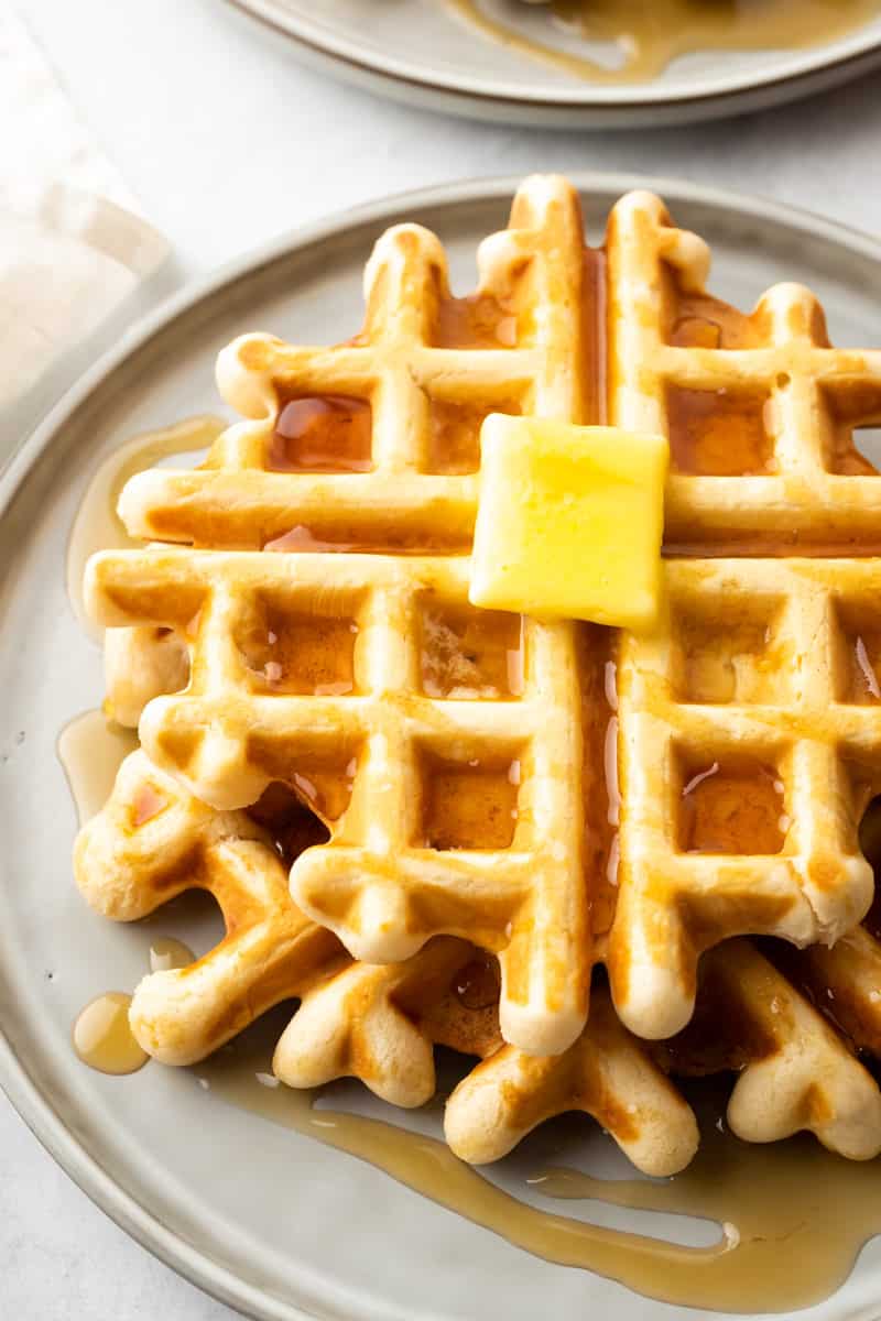 Closeup of a stack of waffles on a beige plate, doused in syrup and topped with a pad of butter