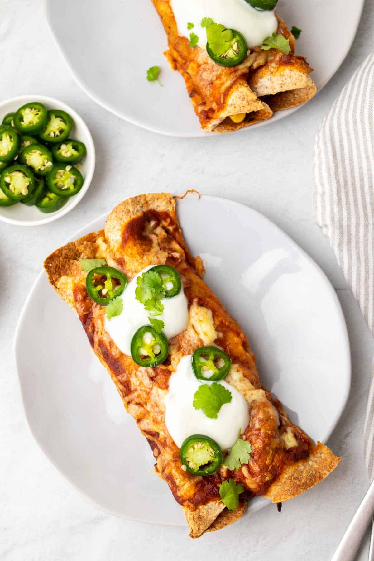 2 enchiladas on a white plate topped with sour cream, sliced jalapenos, and cilantro
