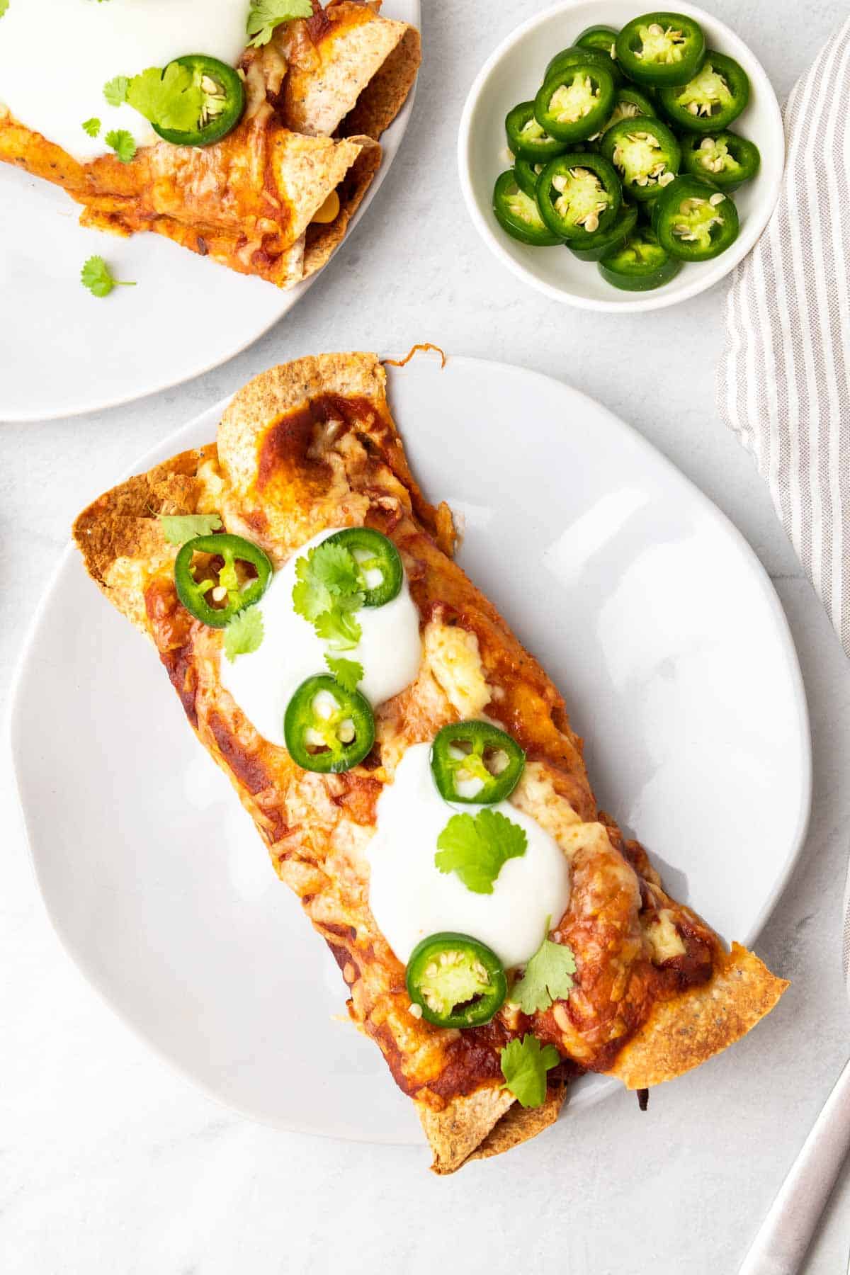 2 enchiladas on a white plate topped with sour cream, sliced jalapenos, and cilantro
