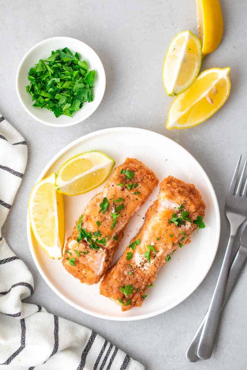 Two filets of baked salmon on a white plate next to two lemon wedges with a ramekin of fresh herbs and more lemon slices
