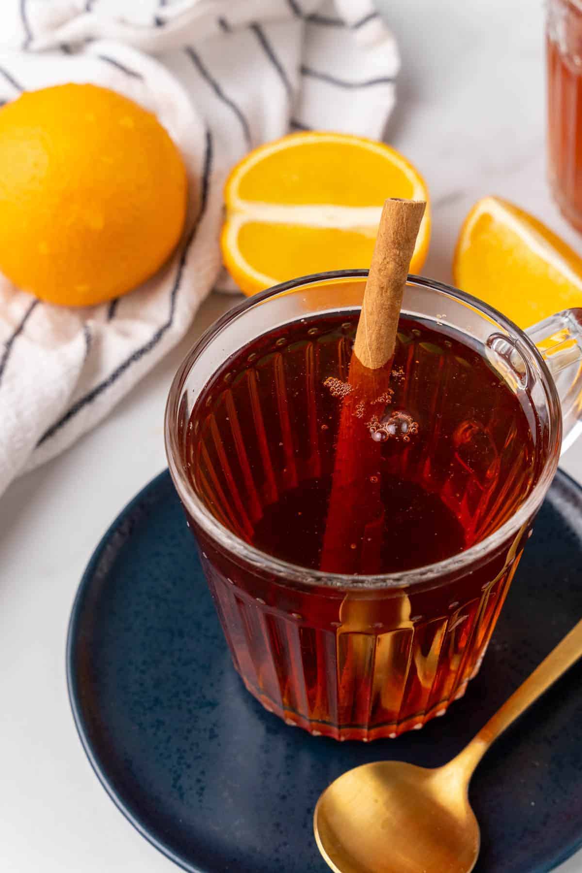 Orange cinnamon breakfast tea in a glass mug with a cinnamon stick on a navy plate with a golden spoon, oranges in the background