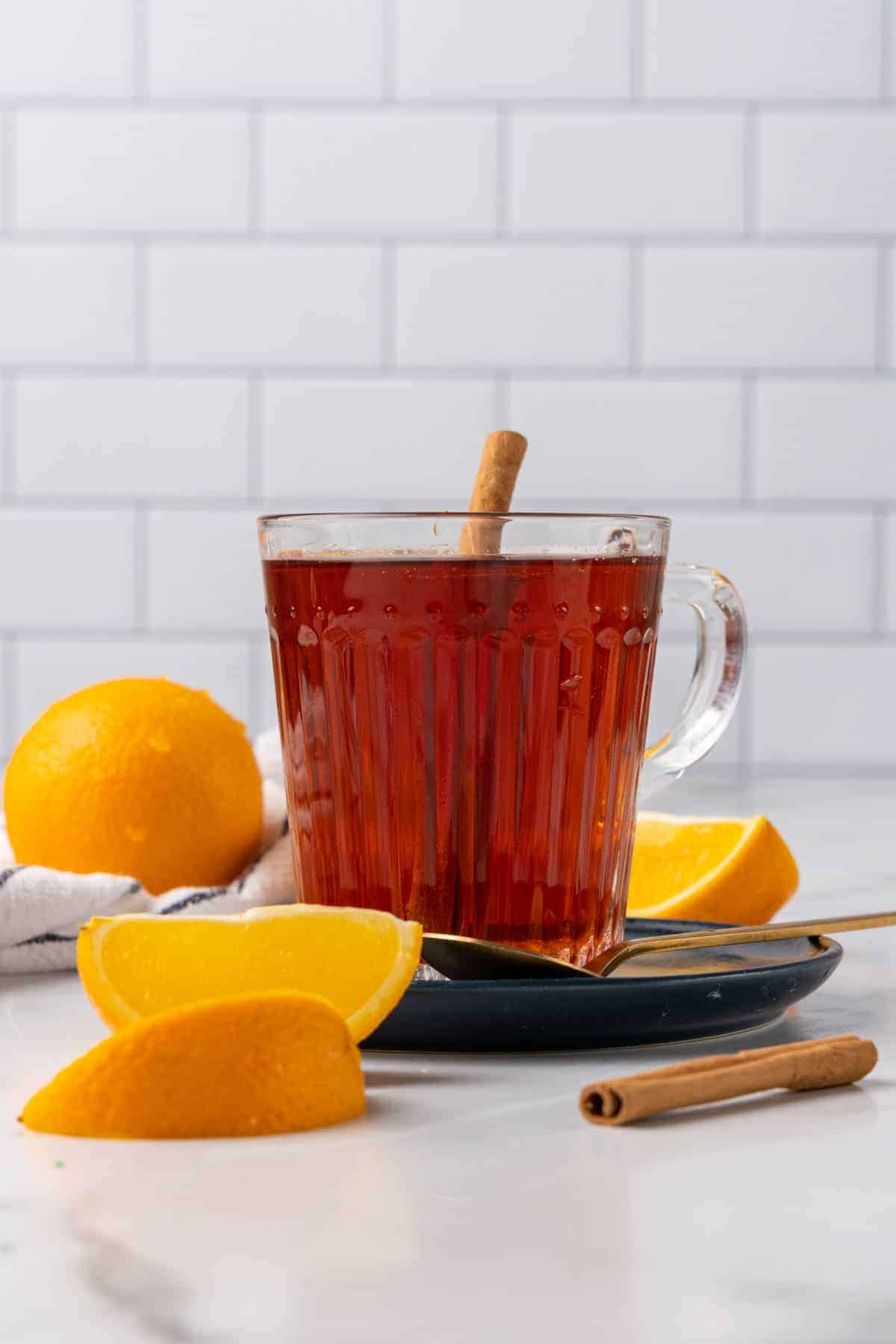 Side view of tea in a glass mug with a cinnamon stick and orange wedges and a cinnamon stick on the counter top around the mug