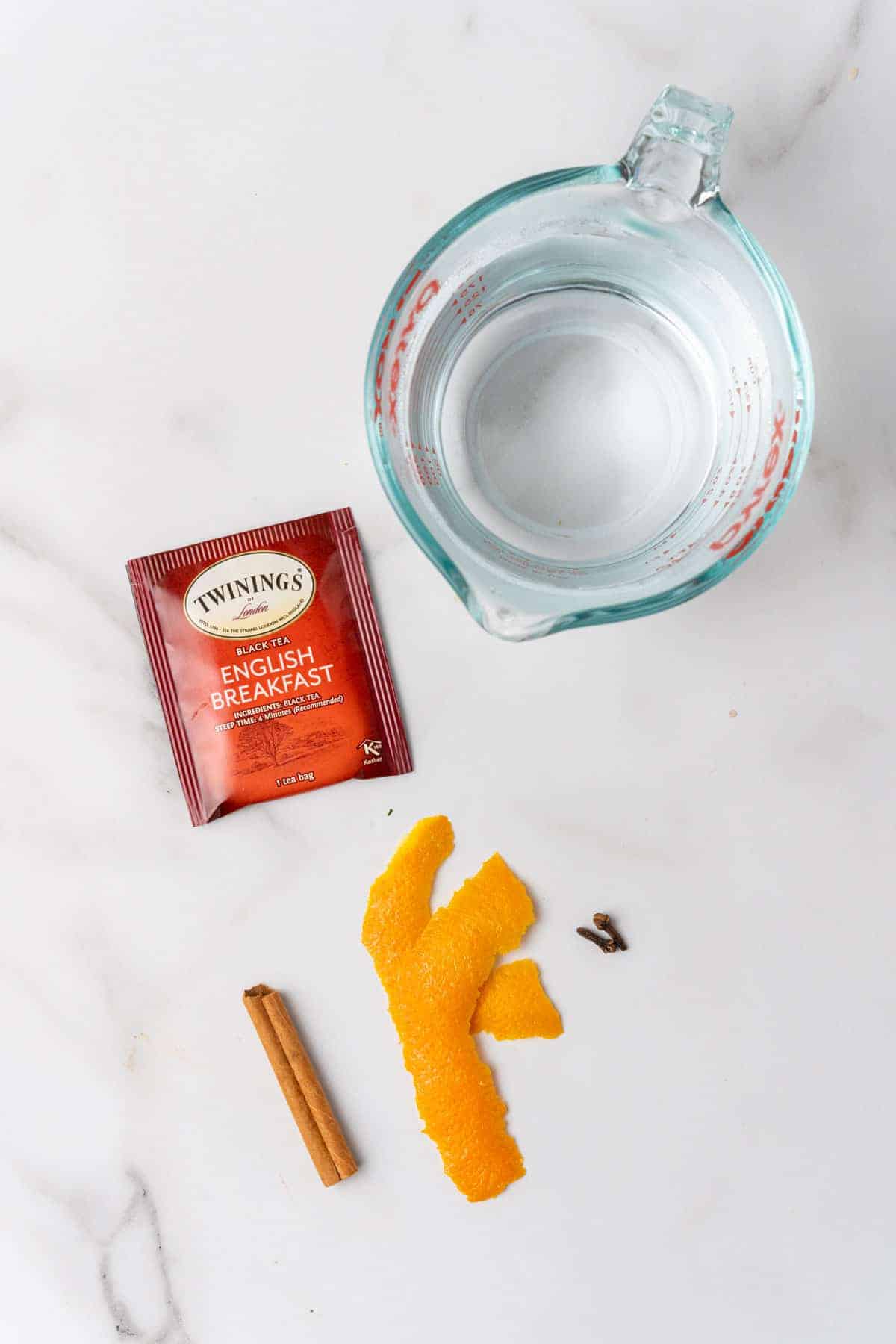 Glass measuring cup, tea bag, cinnamon stick, 2 orange peels, and 2 whole cloves on a white background, as seen from above