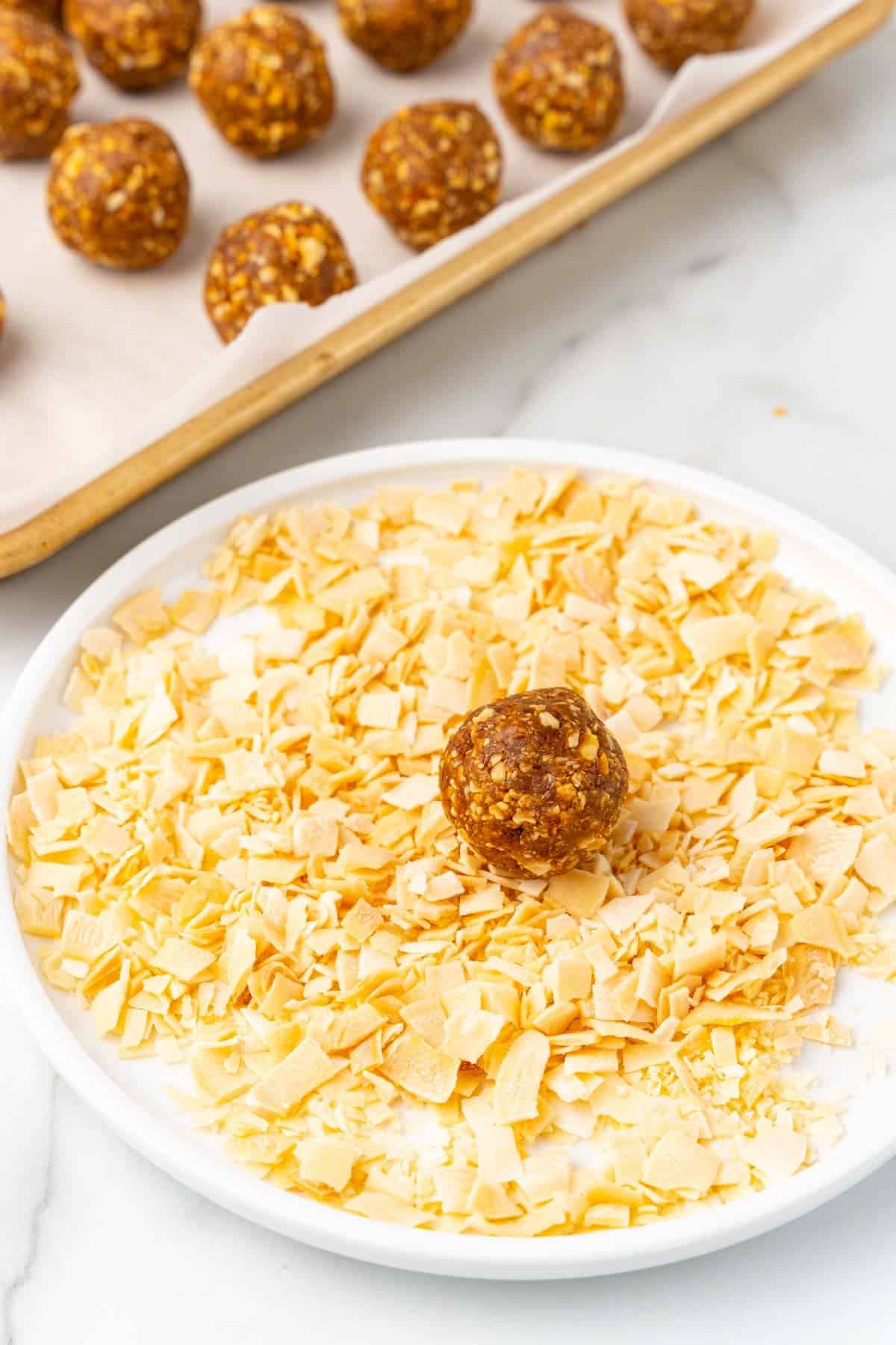One date ball placed on a white plate with chopped toasted coconut; more date balls in the background on a baking sheet lined with parchment paper