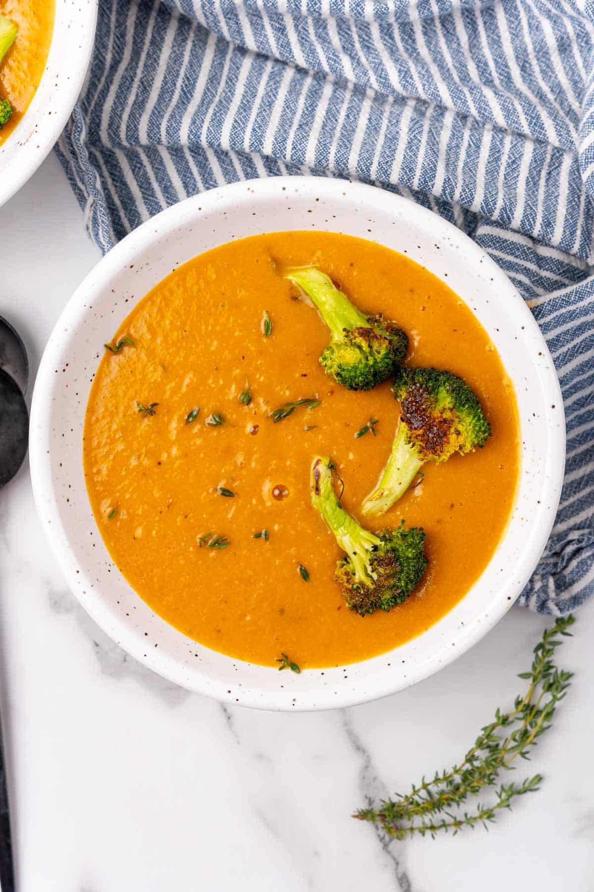 Orange bell pepper soup with roasted broccoli florets on top in a white speckled bowl on a white marble countertop next to a blue striped cloth napkin and a sprig of thyme