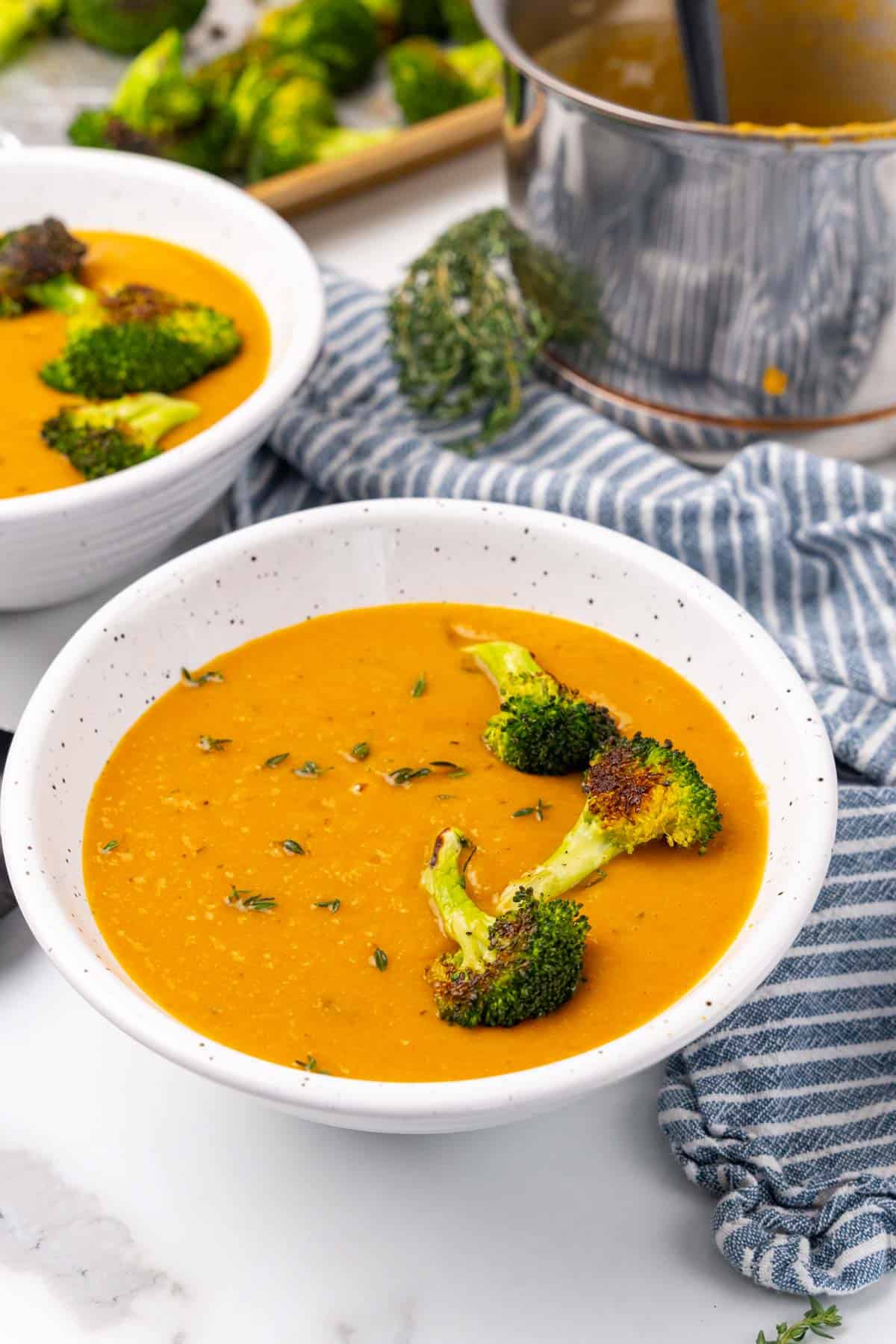 Side view of a white speckled bowl of soup with broccoli florets on top next to a blue striped napkin with a second bowl of soup and saucepan in the background