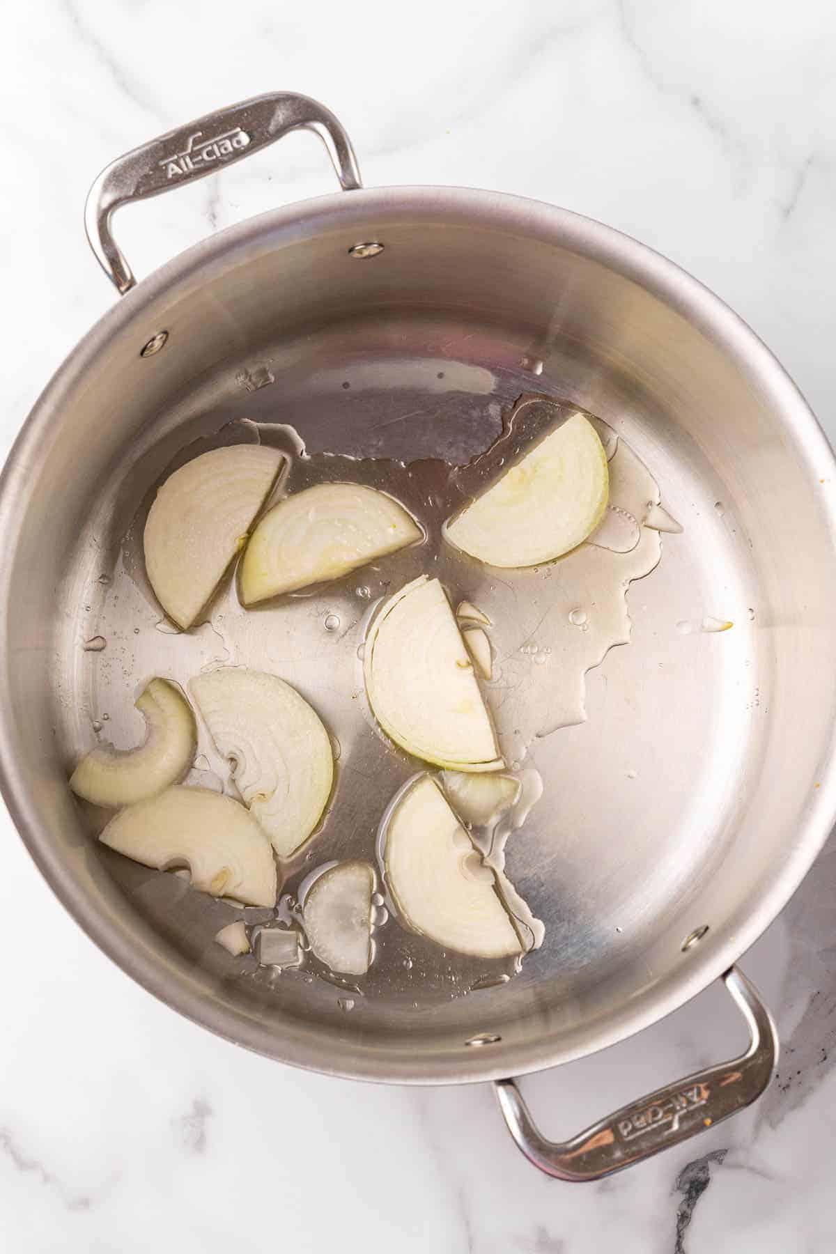 Onions and oil added to a silver saucepan, as seen from above on a white marble countertop