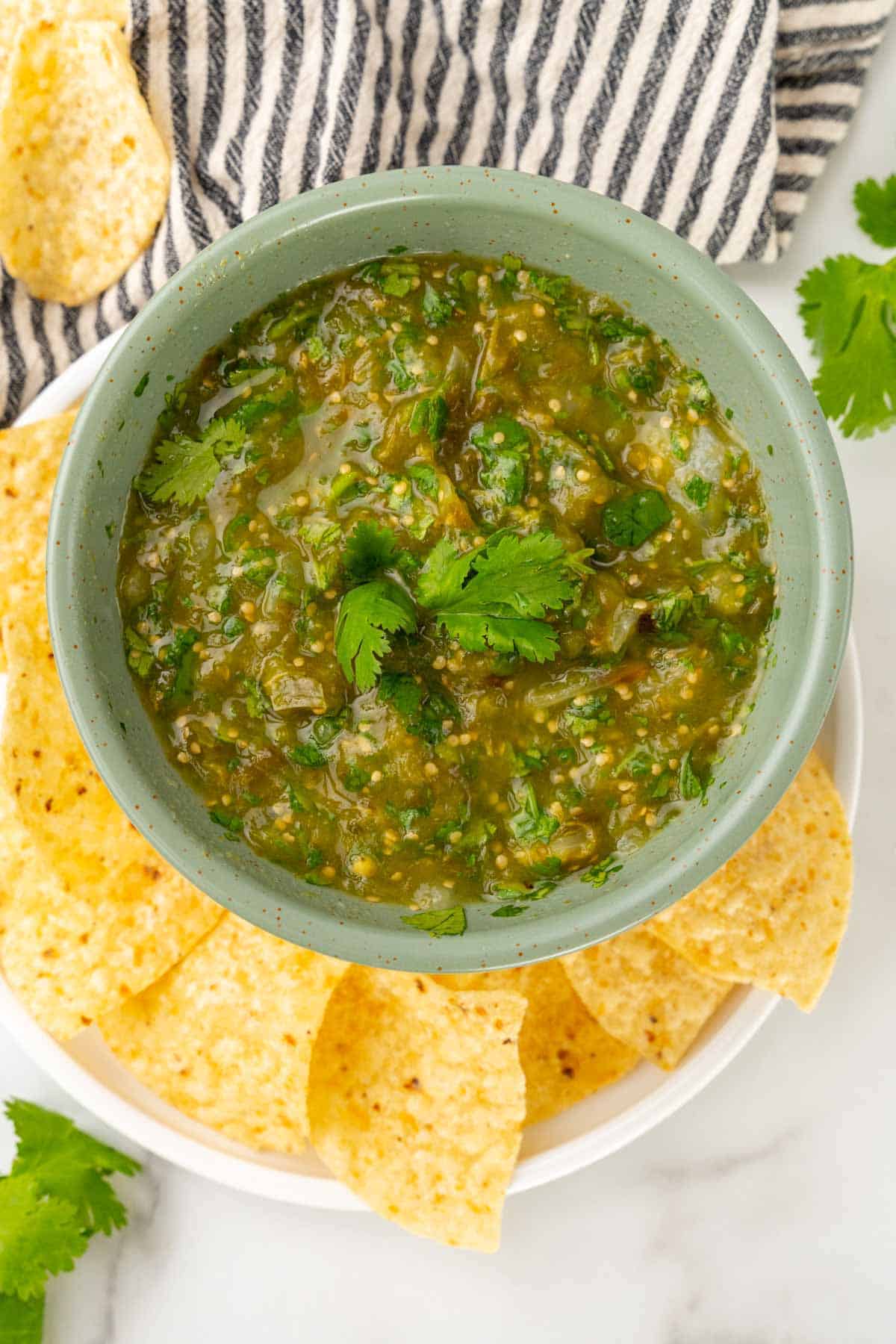 Overhead view of salsa in a light green bowl with cilantro on top; bowl is on a white plate with more tortilla chips on a white marble countertop next to a blue striped cloth napkin with more cilantro and a tortilla chip scattered about
