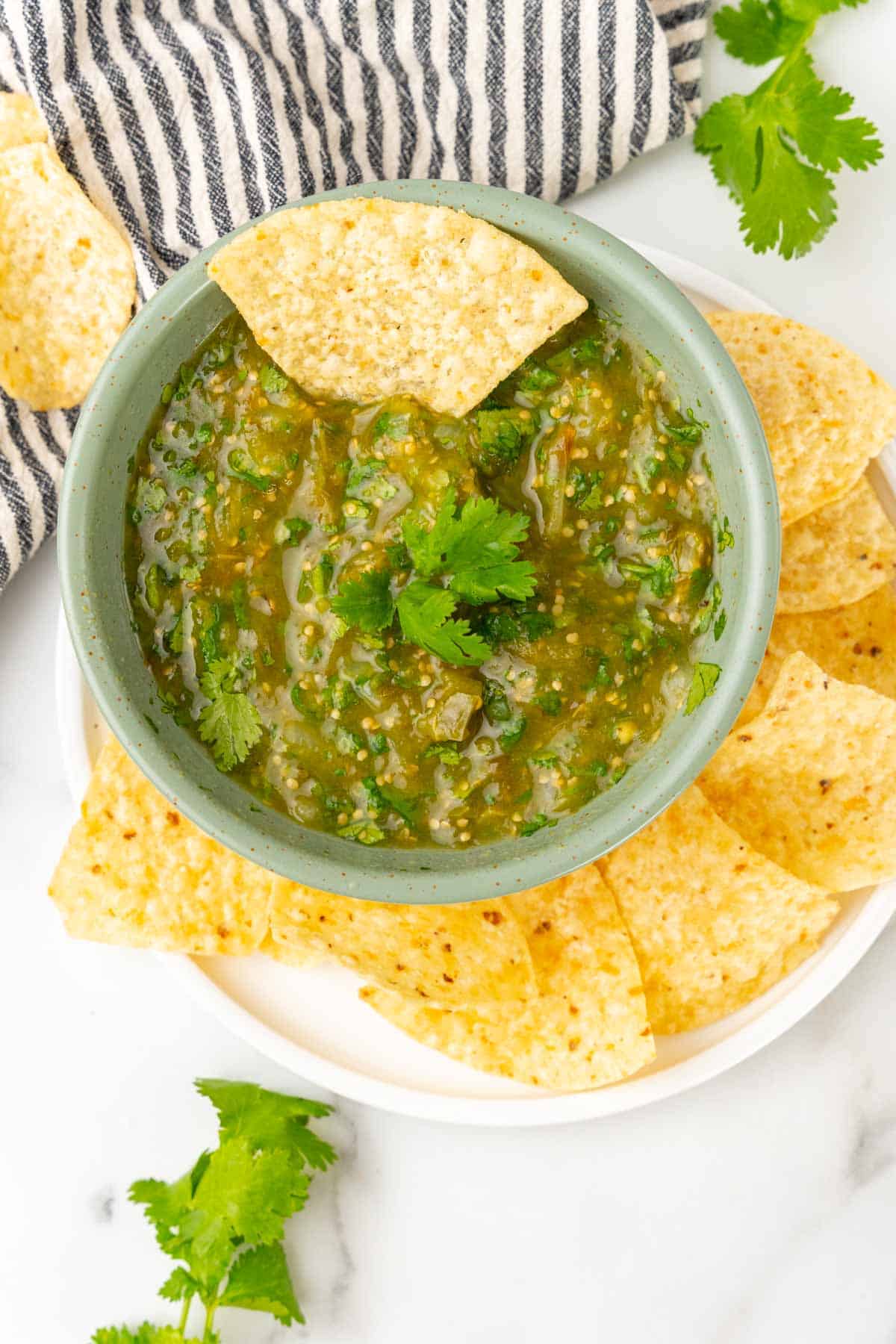 Tomatillo salsa in a light green bowl with cilantro on top and a tortilla chip sticking out of the salsa. Bowl is on a white plate with more tortilla chips on a white marble countertop next to a blue striped cloth napkin with more cilantro and tortilla chips scattered about