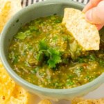 A hand dipping a tortilla chip into the salsa in a green bowl on a white plate surrounded by more tortilla chips with a blue striped cloth napkin in the background