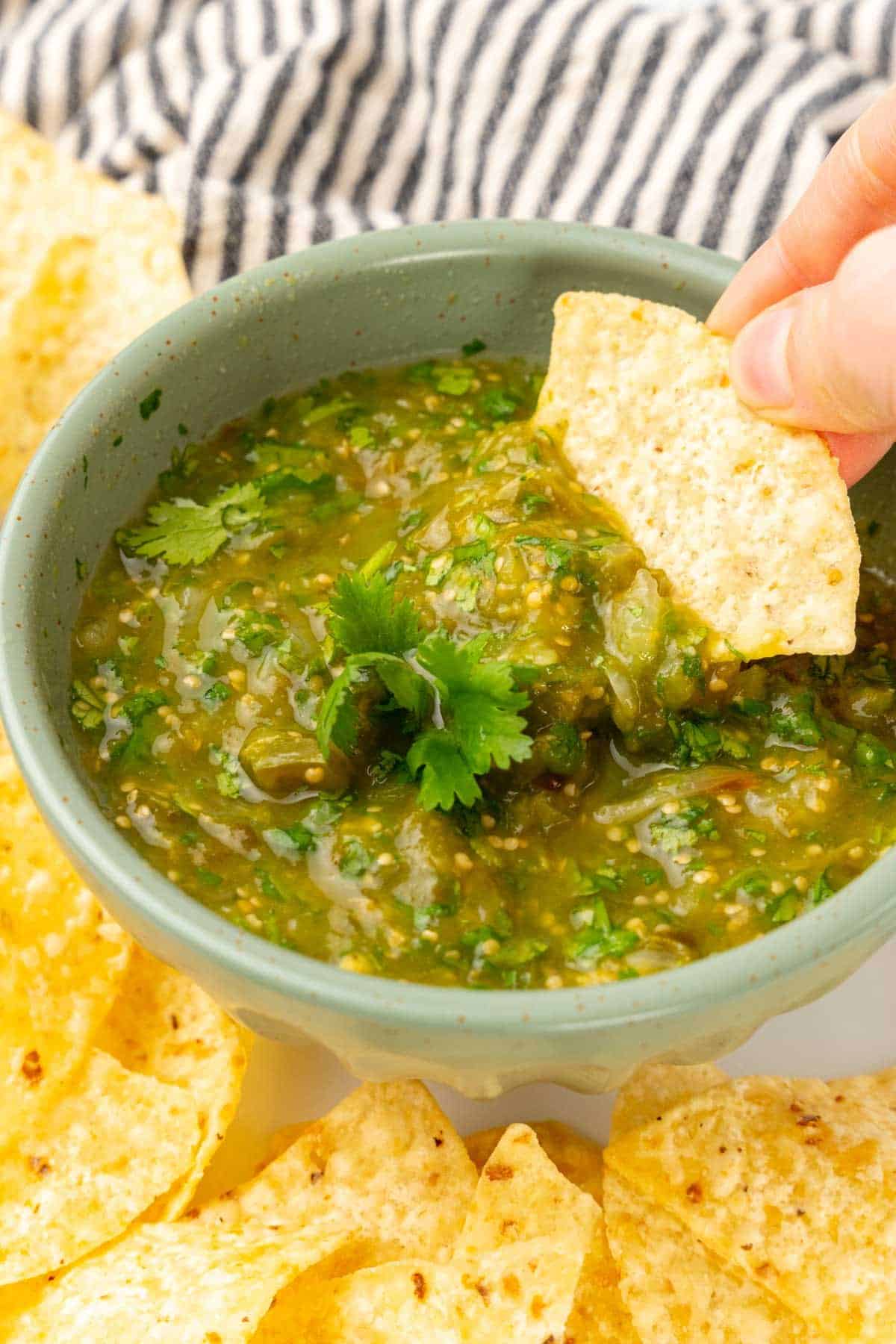 A hand dipping a tortilla chip into the salsa in a green bowl on a white plate surrounded by more tortilla chips with a blue striped cloth napkin in the background