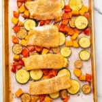 Four roasted salmon filets on top of roasted veggies on a baking sheet lined with parchment paper, as seen from above on a white marble countertop