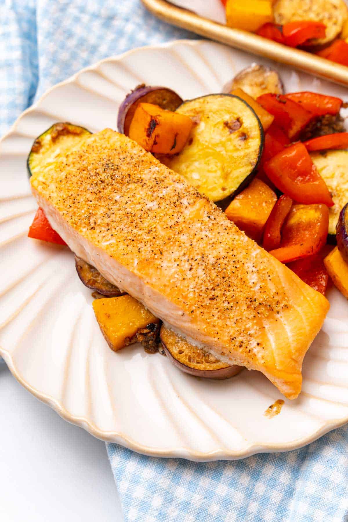 Baked salmon with roasted vegetables on a white plate on a blue checkered cloth napkin