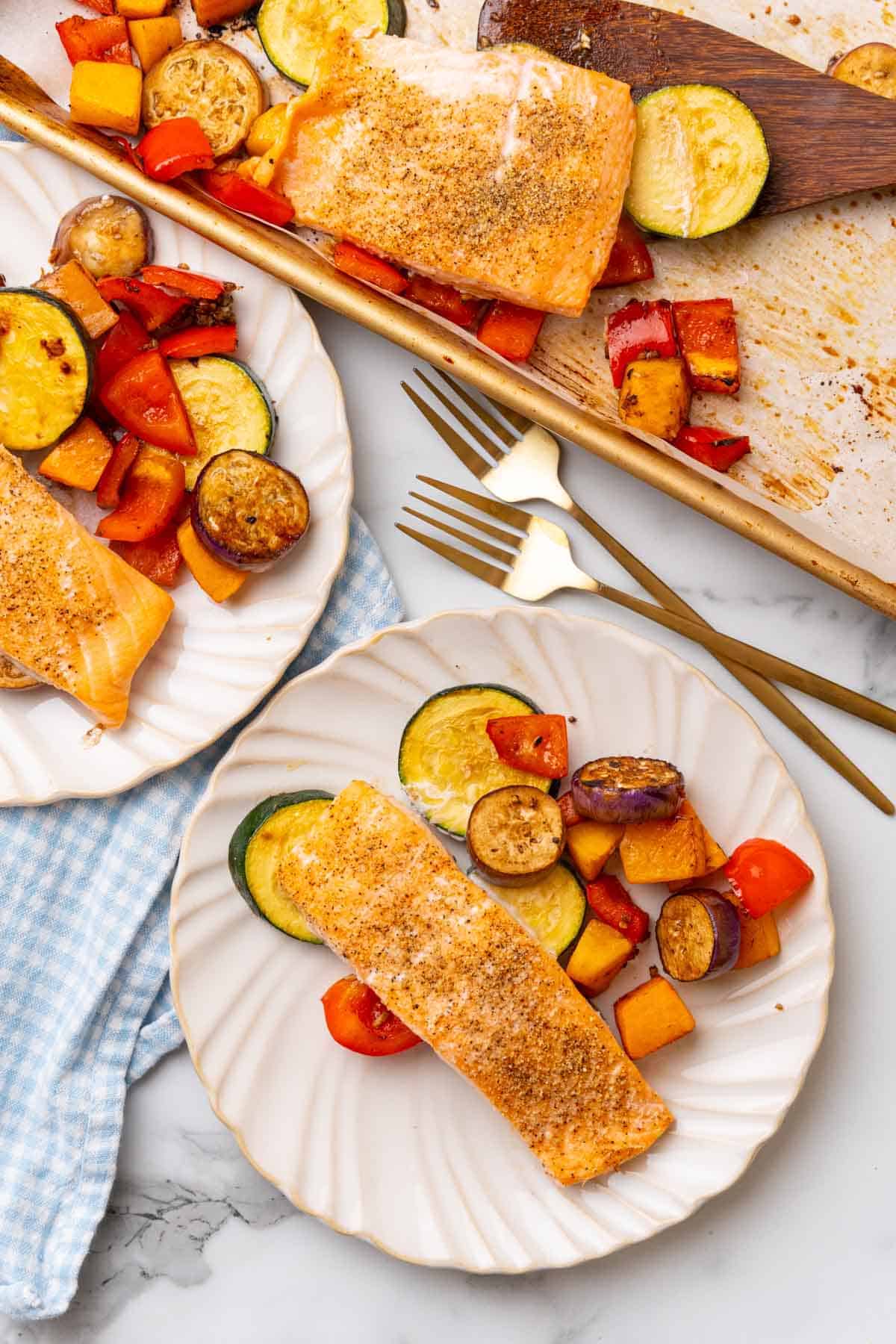 Overhead view of two plates of salmon with roasted veggies next to two gold forks and a baking tray with more salmon and veggies