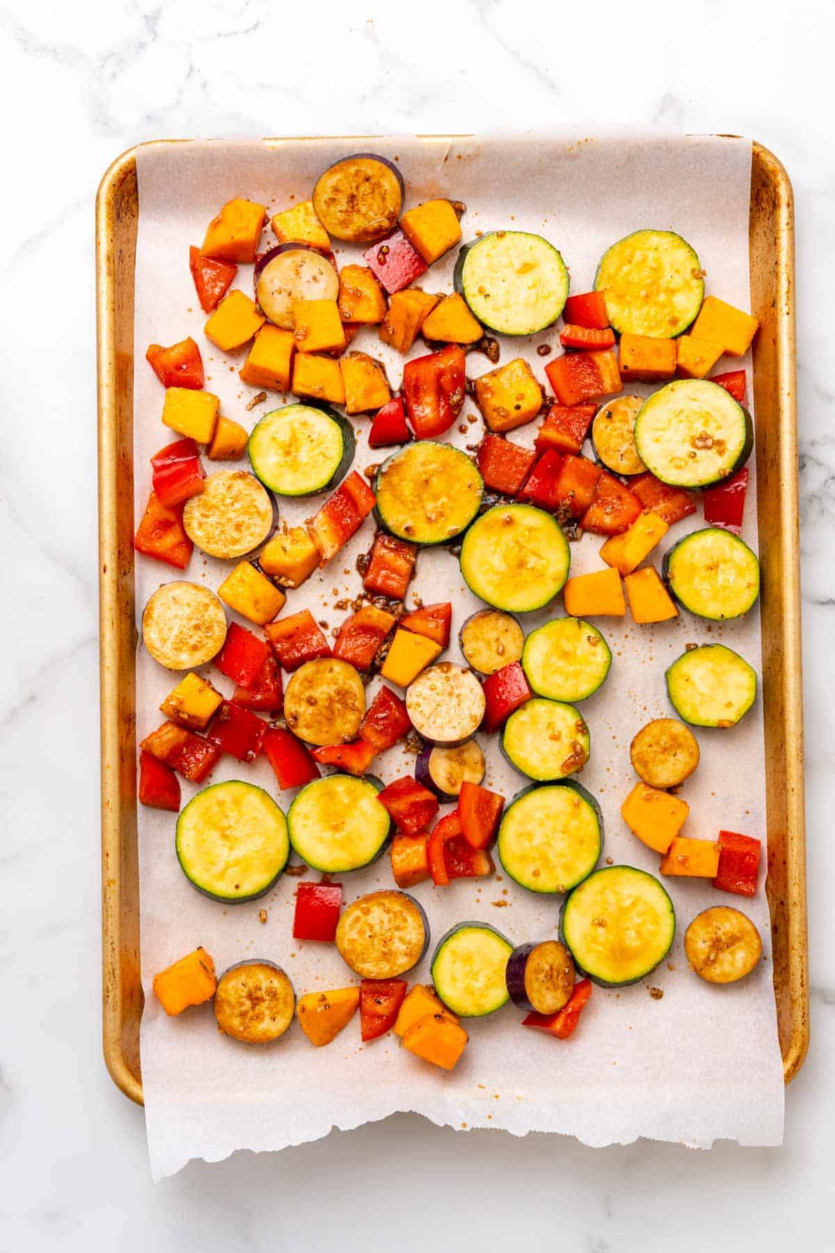 Glazed veggies on a baking tray lined with parchment paper, as seen from above on a white marble countertop
