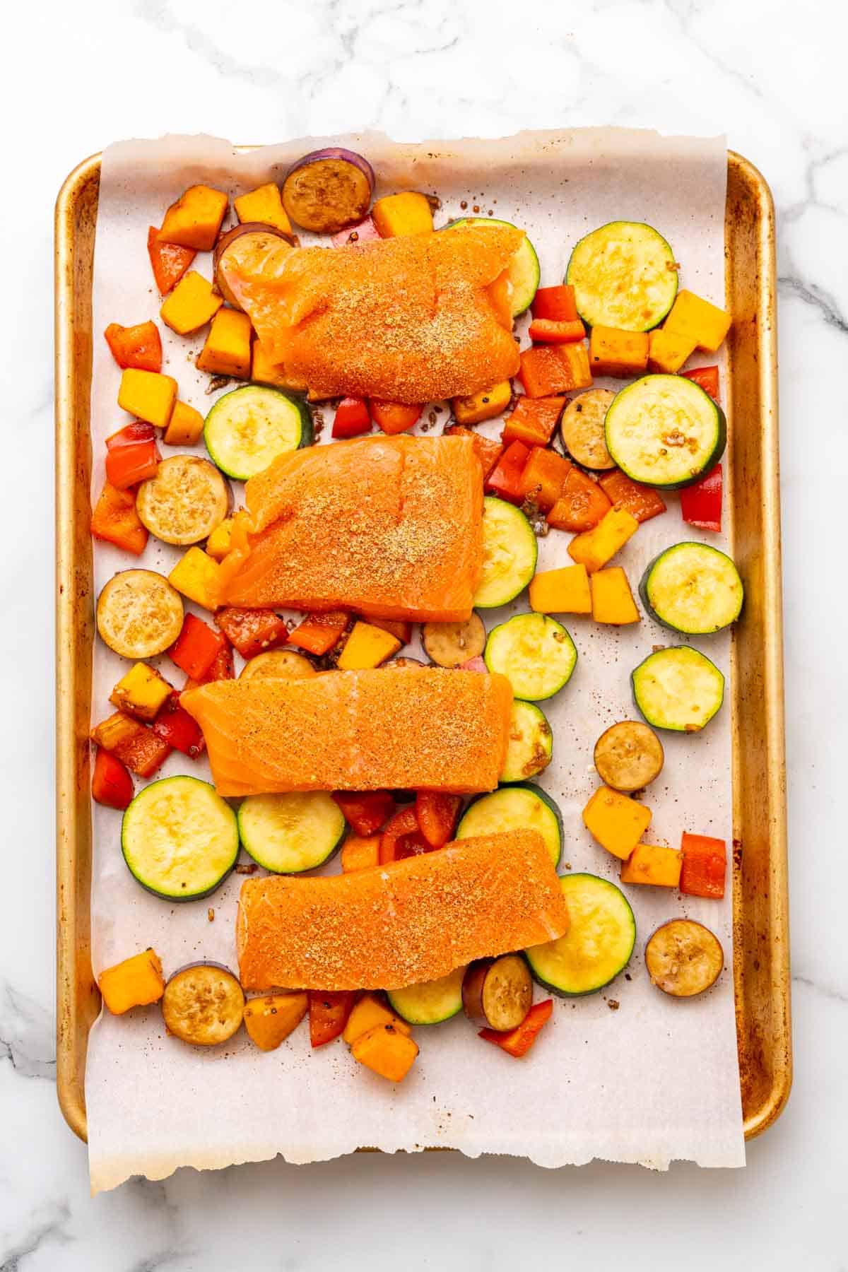 Four salmon filets laid on top of roasted veggies on a baking tray lined with parchment paper, as seen from above on a white marble countertop