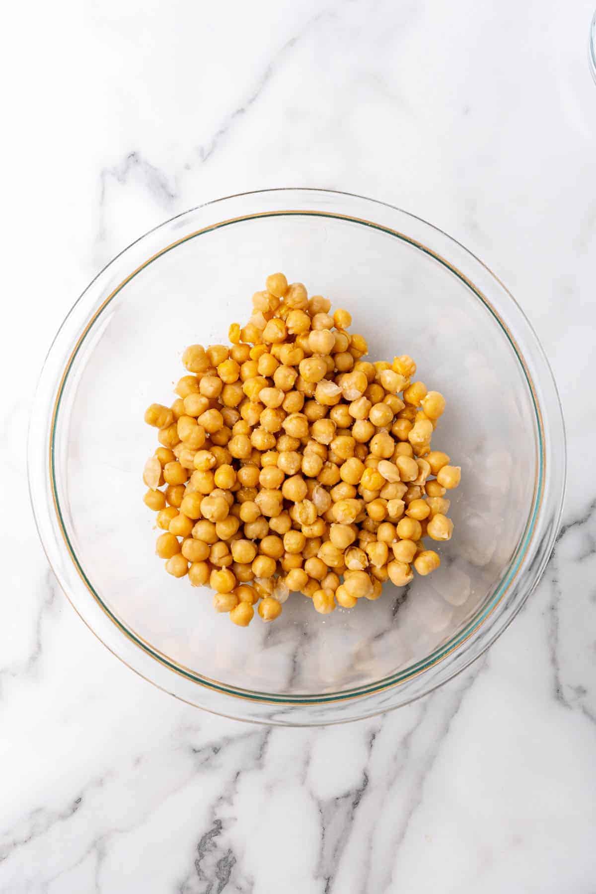 Chickpeas added to a glass bowl, as seen from above on a white marble countertop