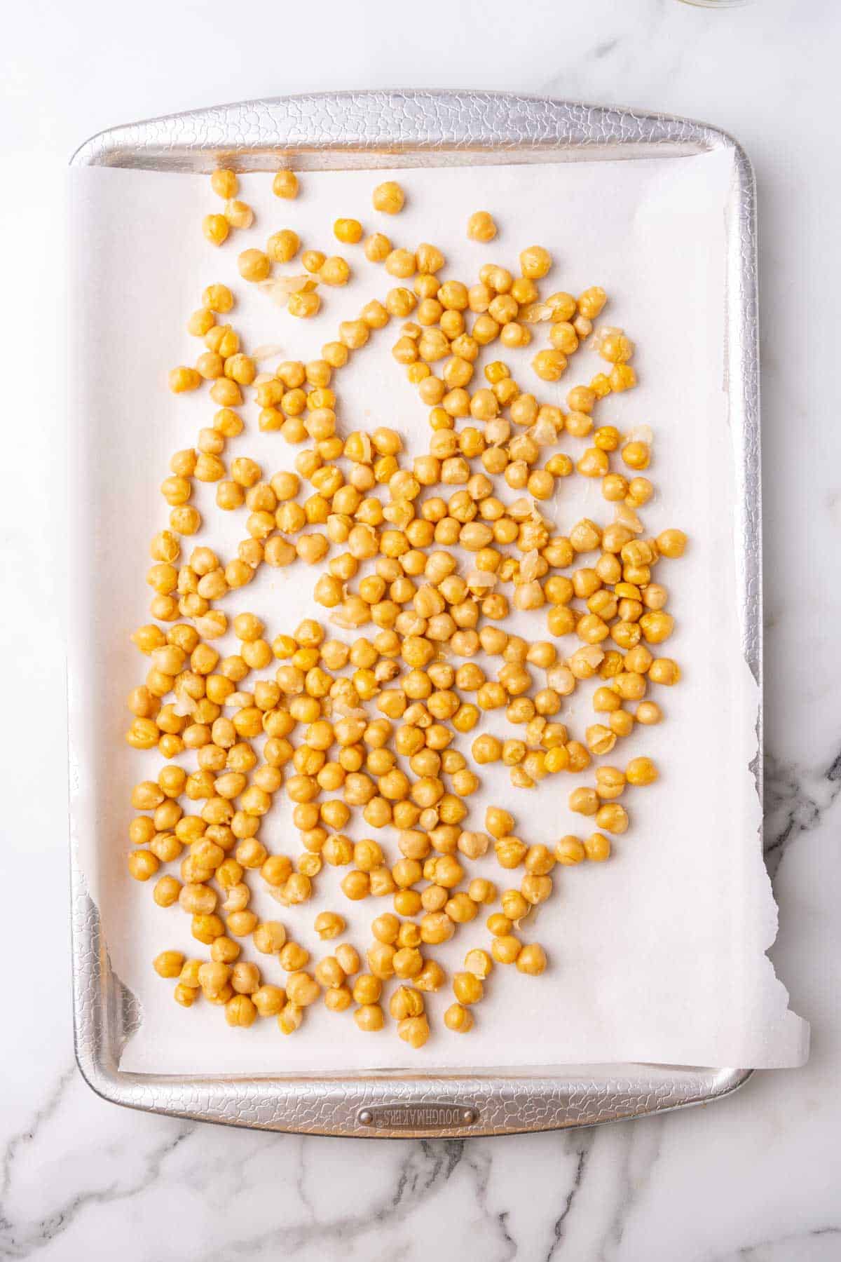 Chickpeas spread out on a baking tray lined with parchment paper, as seen from above on a white marble countertop
