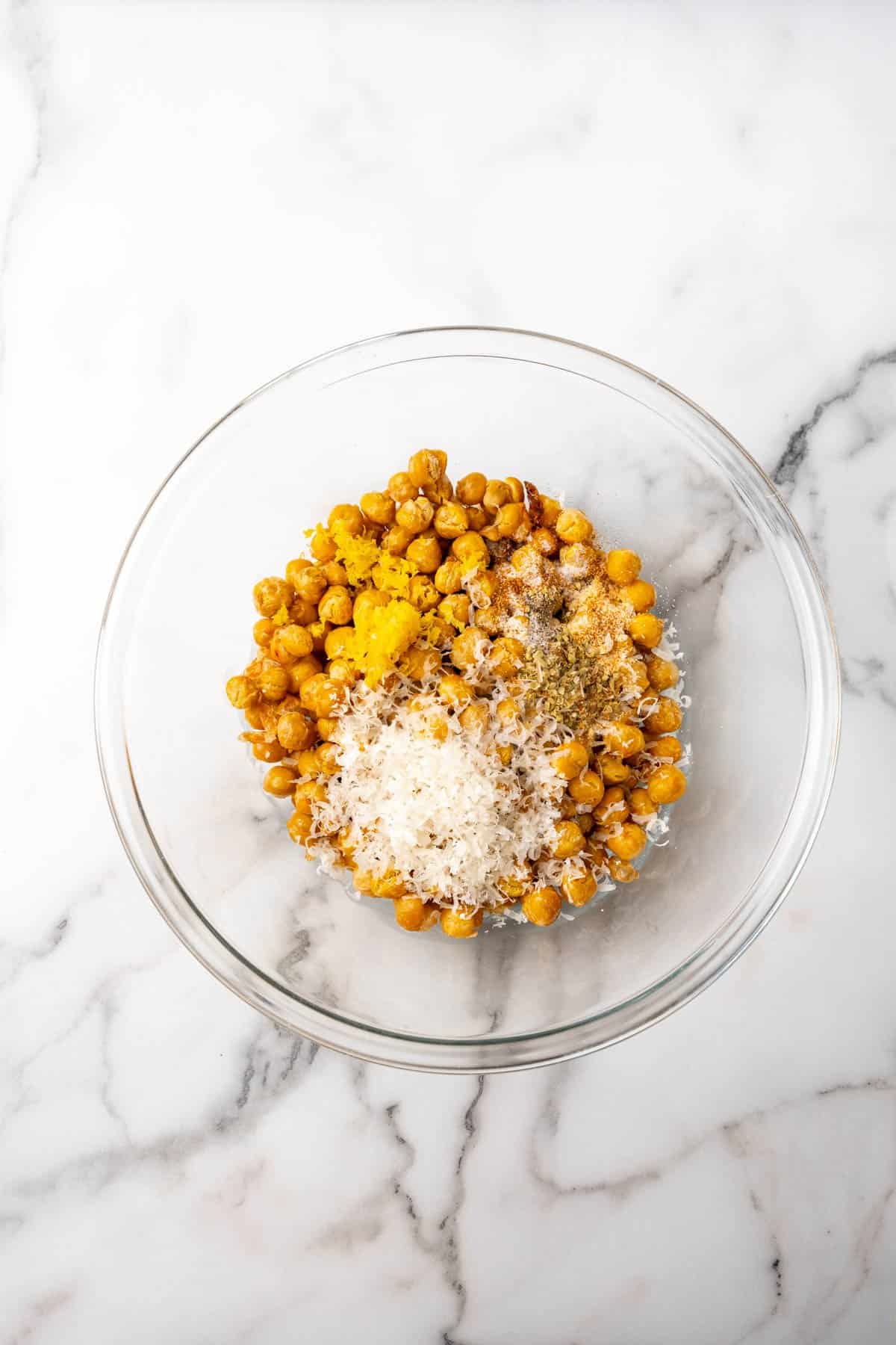 Roasted chickpeas in a glass bowl with parmesan and seasoning added on top, as seen from above on a white marble countertop