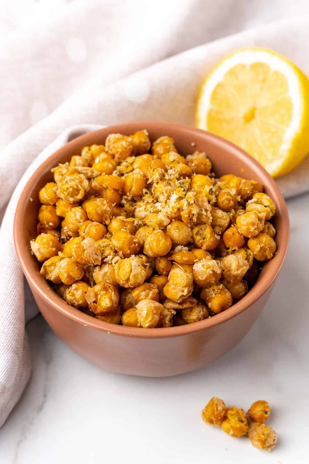 Crispy roasted chickpeas in an orange bowl with an off-white cloth napkin and half of a lemon in the background