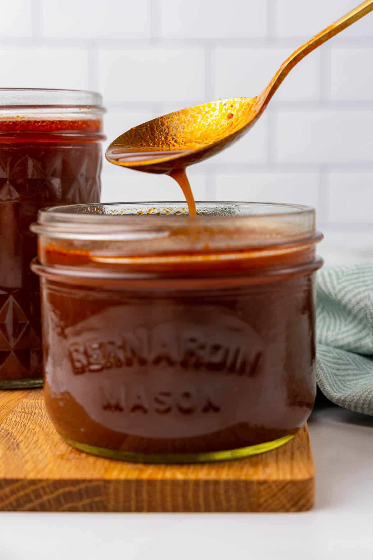 Closeup of brown sauce in a short mason jar on a wooden serving tray with a gold spoon dripping sauce back into the jar